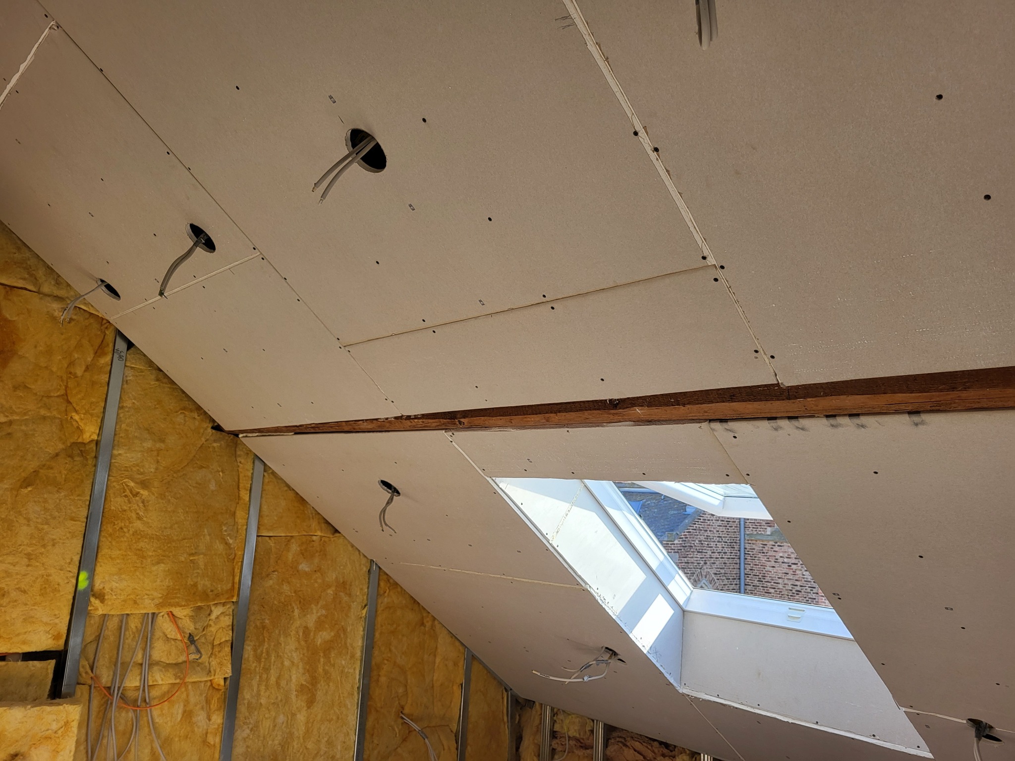 Poddasze w trakcie adaptacji: ocieplone ściany wełną mineralną, zamontowana płyta gipsowo-kartonowa na skosach sufitu z wyciętymi otworami na oświetlenie i oknem dachowym.