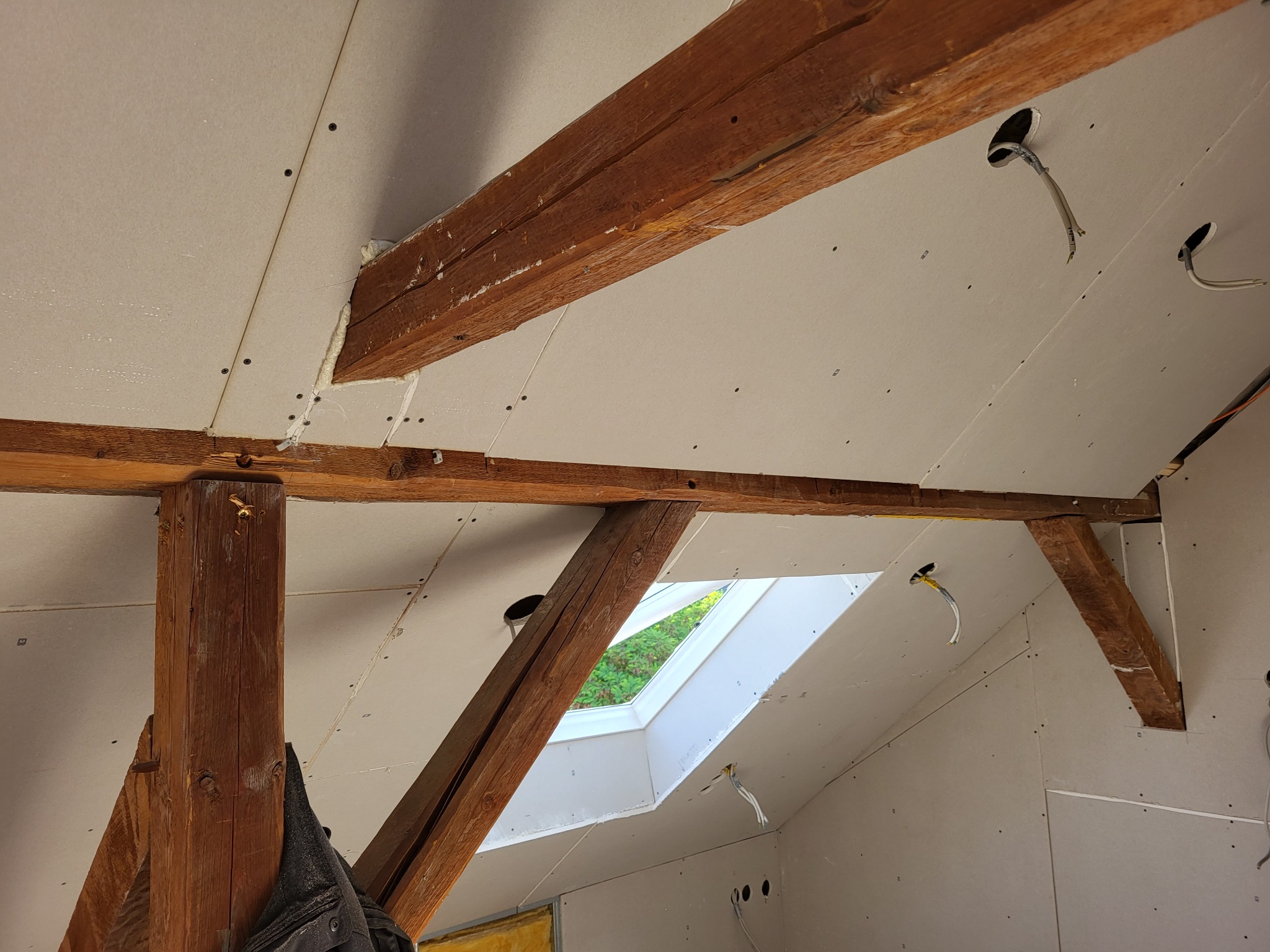 Poddasze w trakcie wykańczania: widoczne drewniane belki konstrukcyjne, płyty gipsowo-kartonowe zamontowane na skosach, okno dachowe oraz otwory na instalacje elektryczne z wystającymi przewodami.