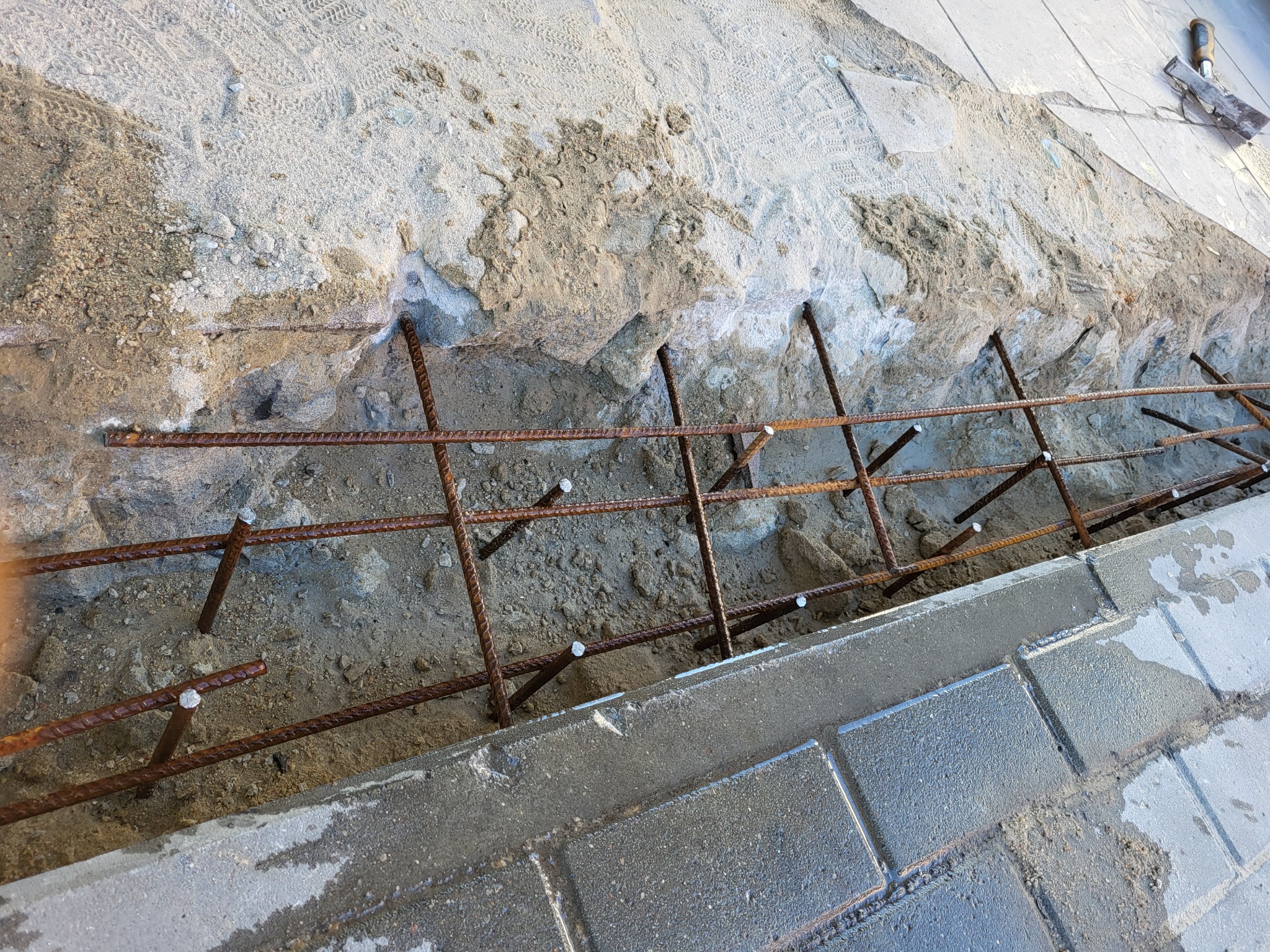 Zbrojenie z prętów stalowych w wykopie, przygotowane do zalania betonem pod obrzeże z kostki brukowej. W tle widoczna kielnia.