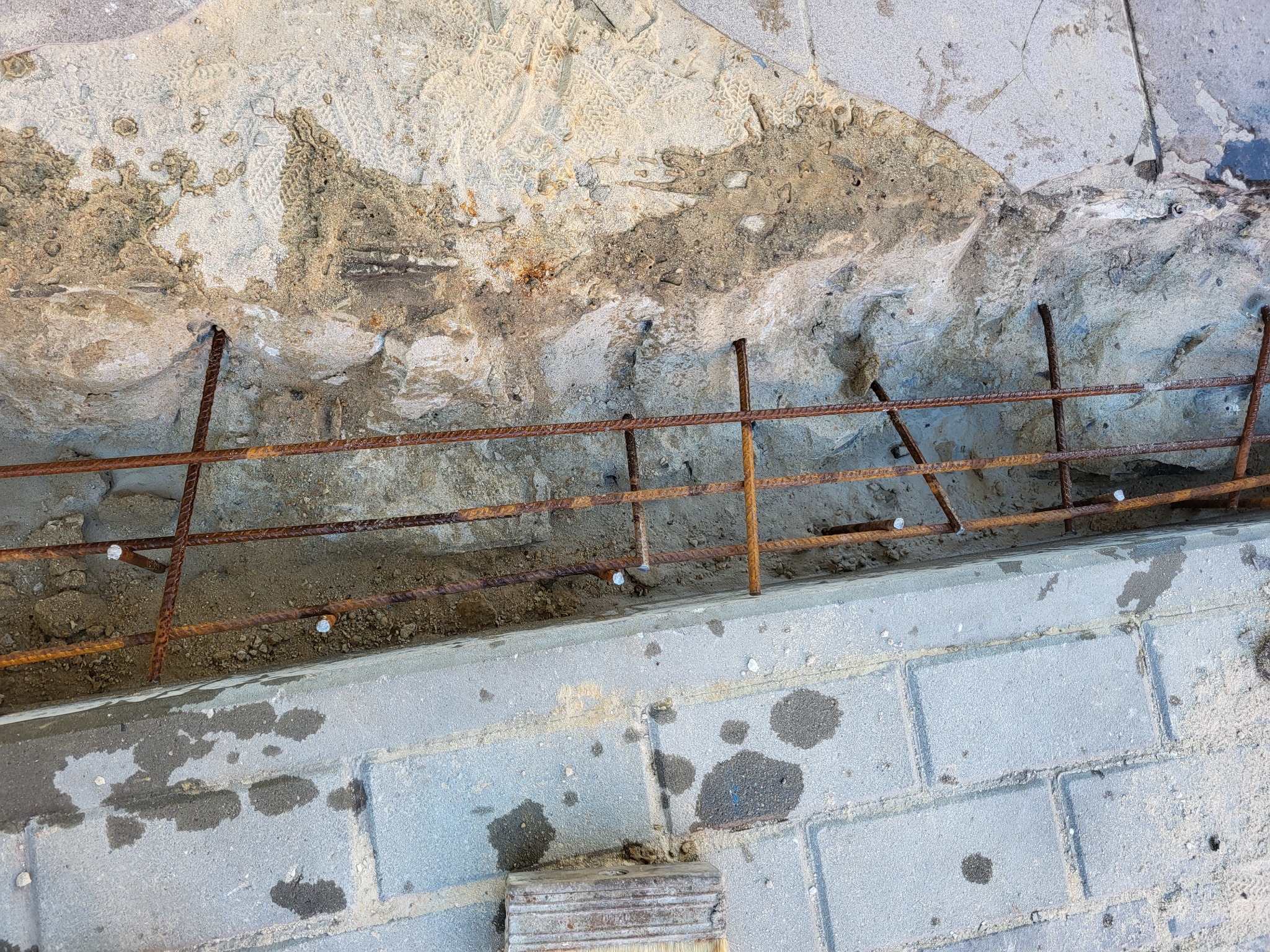 Zbrojenie wystające ze ściany nad świeżo ułożoną kostką brukową, widoczne ślady butów w zaprawie.