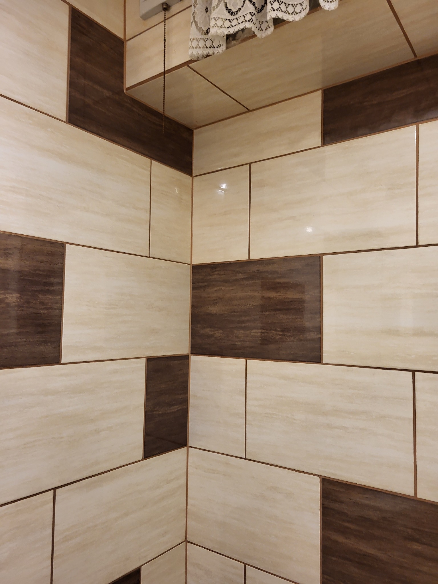 Wykończona ściana w łazience z beżowych i brązowych płytek imitujących marmur i drewno, ułożonych w prostokątnym wzorze. Widoczny fragment sufitu z dekoracyjną koronką.