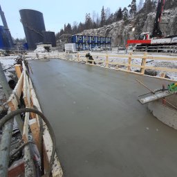 CBT BUILDING - Świeżo wylany beton na budowie, szalunek z drewnianych desek, w tle zbiorniki i żuraw budowlany.