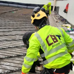 Pracownicy w odblaskowych ubraniach układają siatkę zbrojeniową na budowie przemysłowej, widoczny budynek w tle.