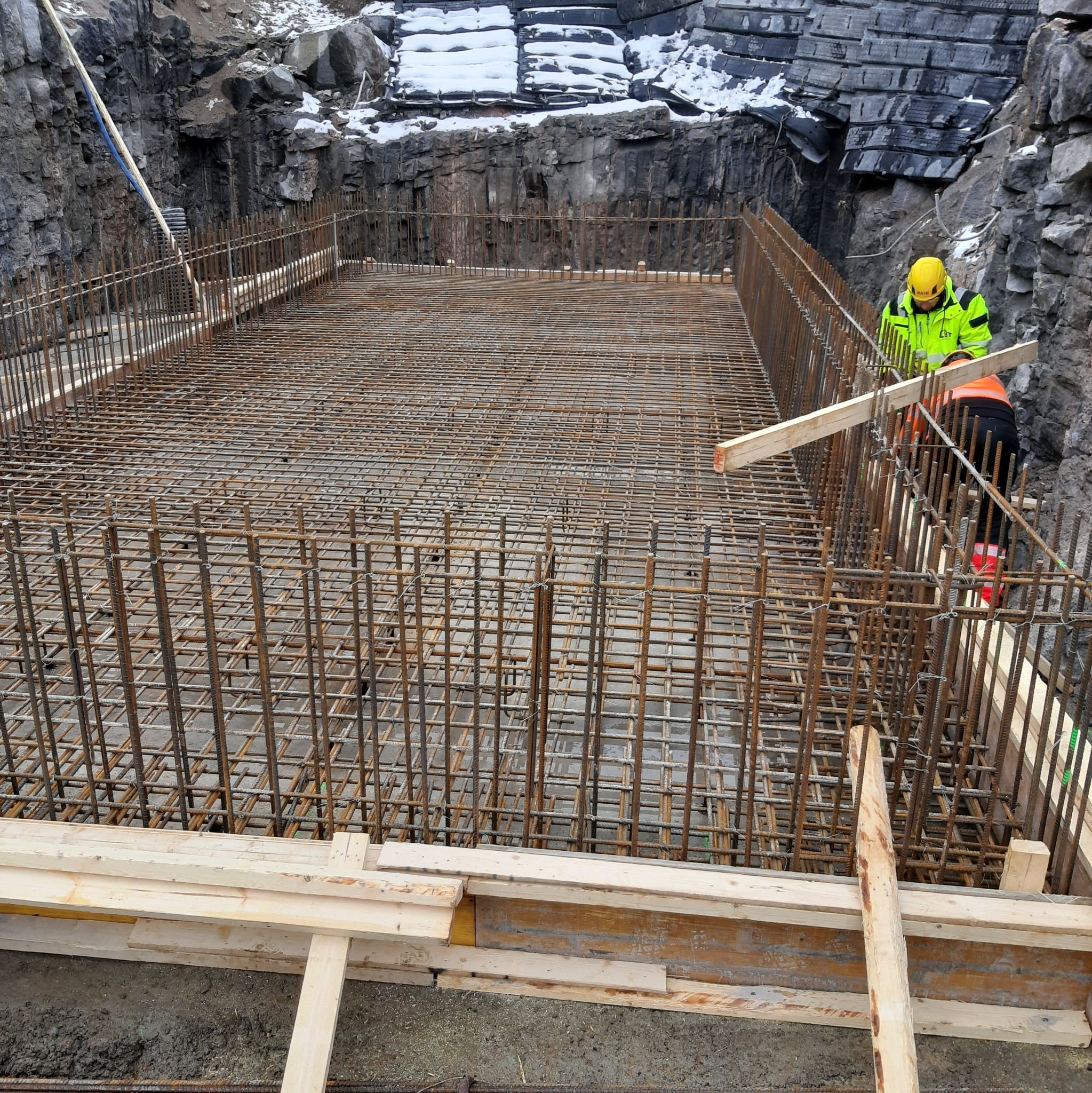 Wykonywanie zbrojenia płyty fundamentowej w Poznaniu, widoczne stalowe pręty tworzące siatkę, drewniane szalunki i pracownik w odblaskowej kurtce na tle skalistego wykopu.