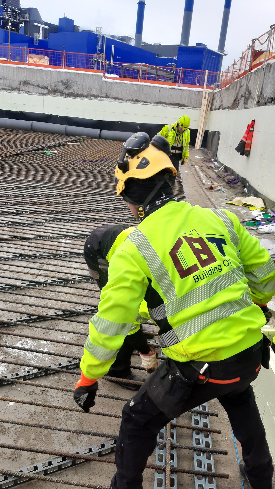 Pracownicy w odblaskowych ubraniach układają siatkę zbrojeniową na budowie przemysłowej, widoczny budynek w tle.