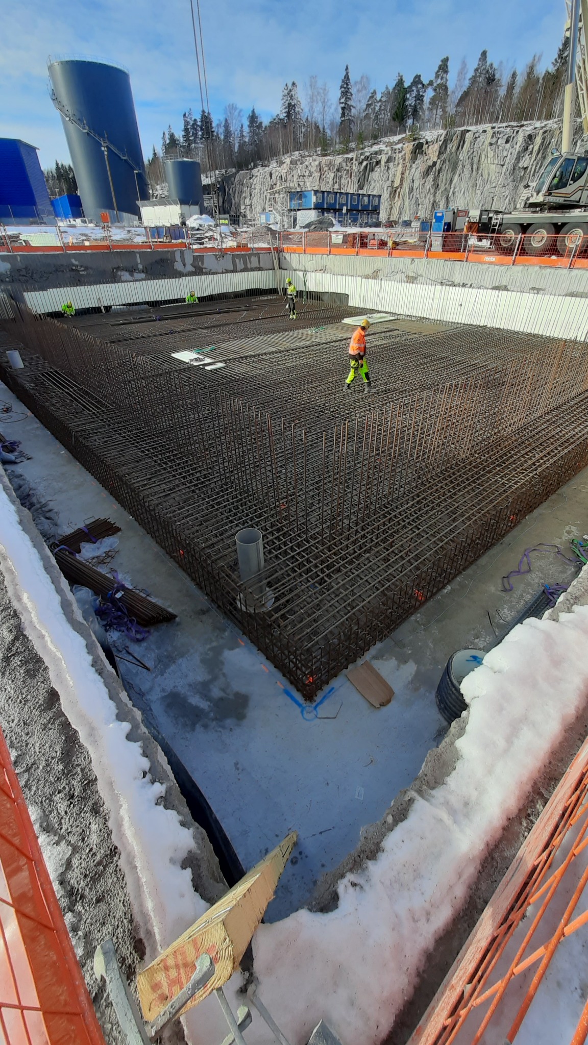 Siatka zbrojeniowa ułożona w wykopie fundamentowym, pracownicy w odblaskowych ubraniach, w tle zbiorniki przemysłowe i skaliste zbocze.