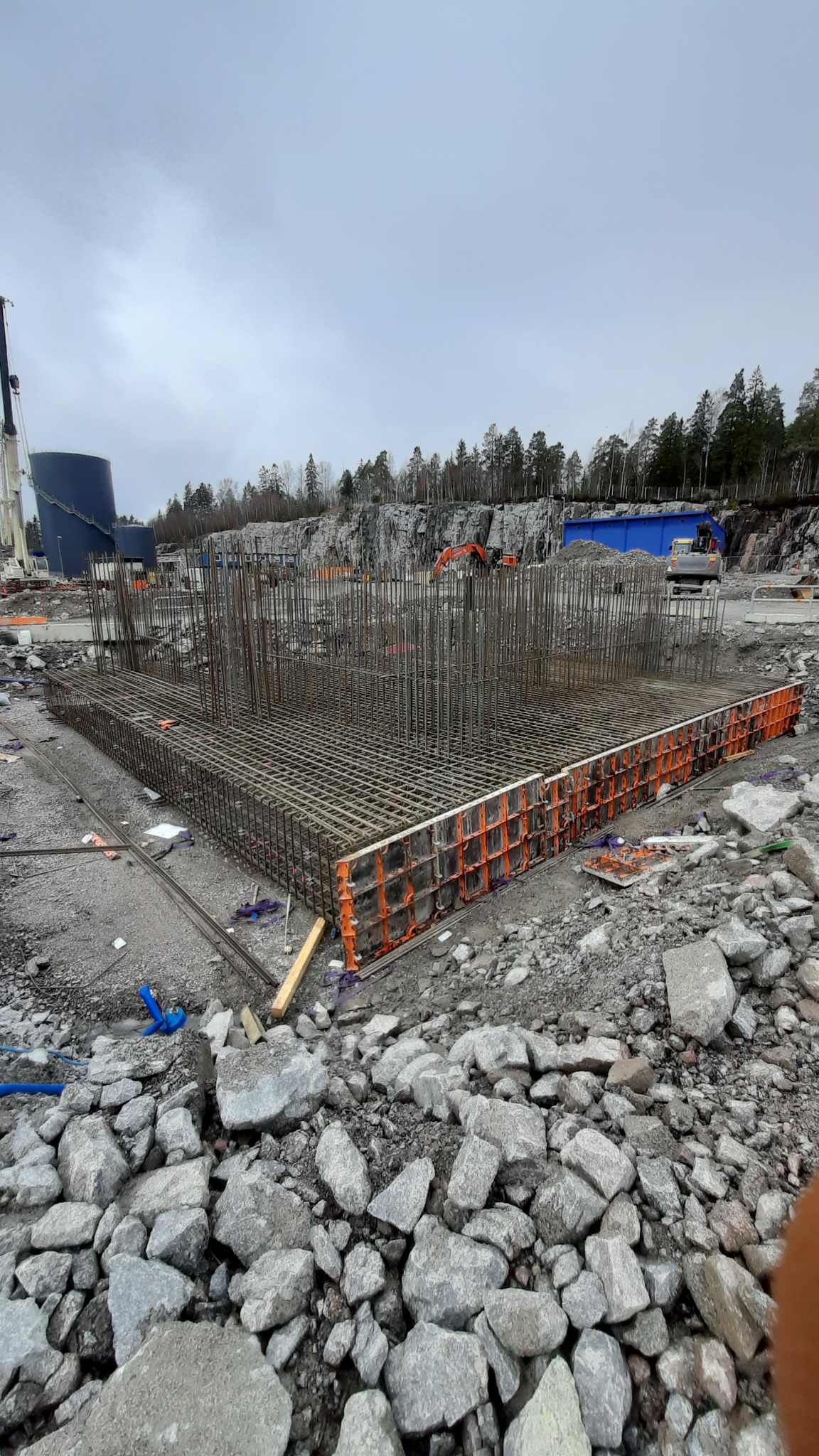 Zbrojenie fundamentu z szalunkiem systemowym na placu budowy, w tle skały i zbiorniki przemysłowe.