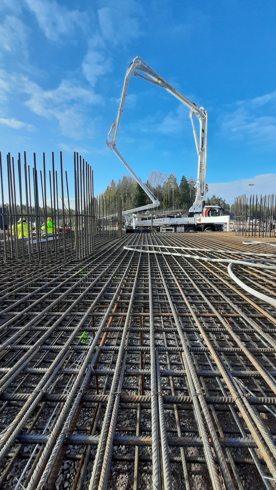 Siatka zbrojeniowa na żwirowym podłożu, widoczny wysięgnik pompy do betonu i pracownicy w odblaskowych kamizelkach w tle.
