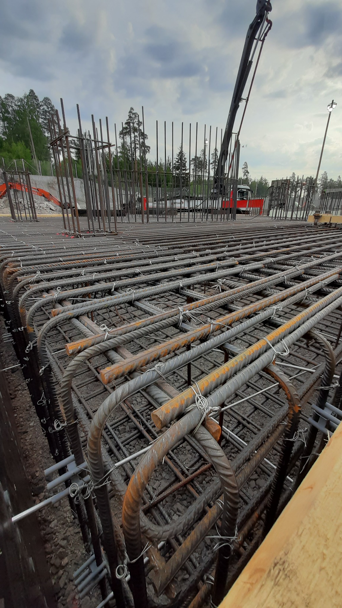 Siatka zbrojeniowa fundamentu z widocznymi prętami pionowymi i podkładkami dystansowymi, w tle maszyna budowlana i drzewa.