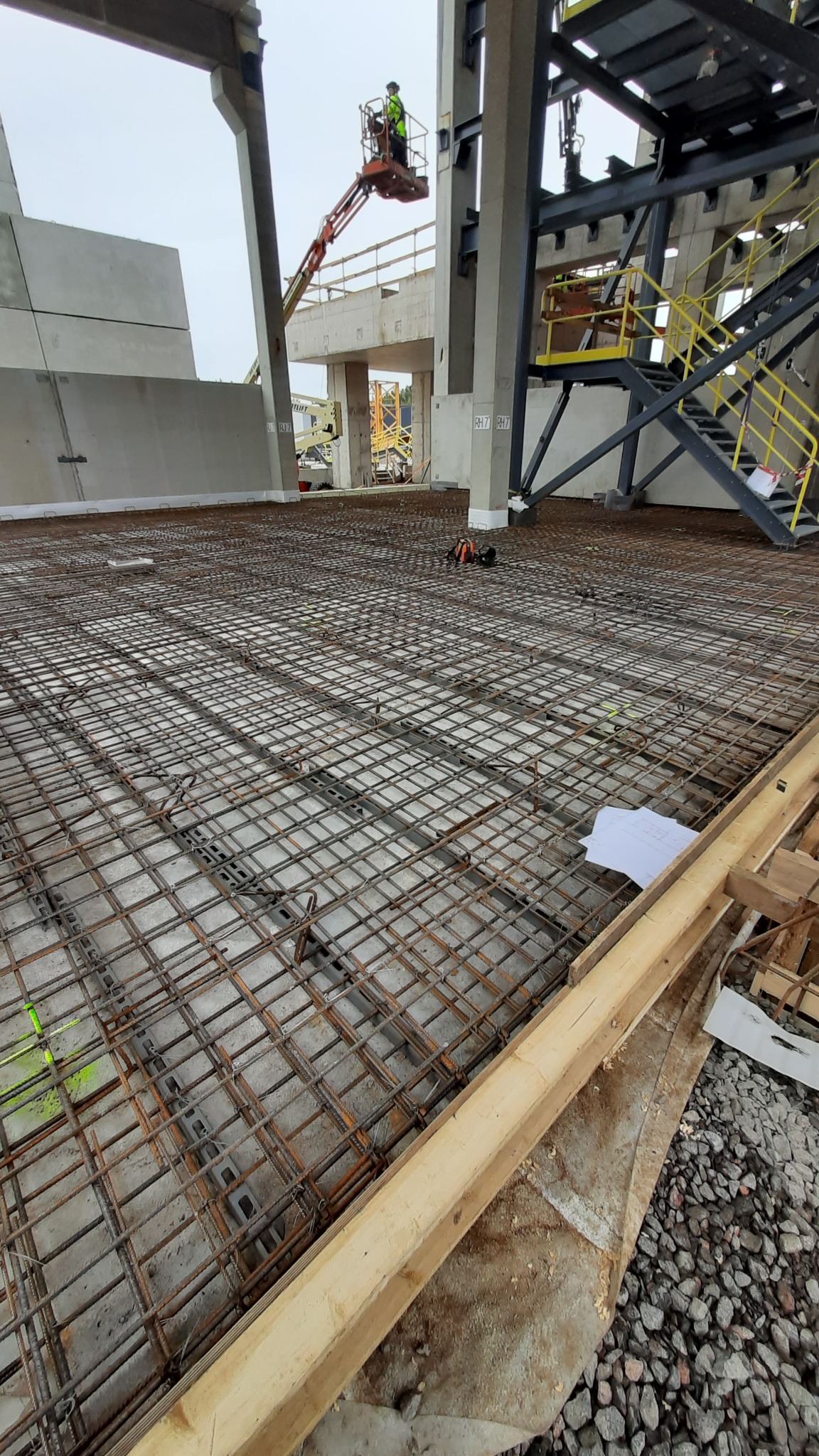 Siatka zbrojeniowa ułożona na betonowej płycie, widoczny pracownik na podnośniku koszowym w tle, elementy konstrukcji stalowej i betonowej.