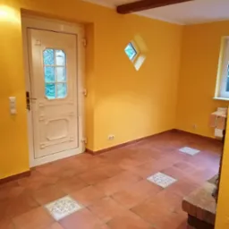 Pomarańczowy pokój z terakotową podłogą, w której wkomponowane są dekoracyjne płytki, oraz z białymi drzwiami i małym romboidalnym oknem w ścianie.