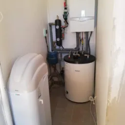 Kotłownia w trakcie instalacji: biały piec gazowy Vaillant, zbiornik na wodę, zmiękczacz wody Primo USTM, manometry, rury i armatura w pomieszczeniu z szarą podłogą.