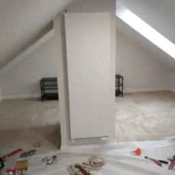 Biały grzejnik panelowy zamontowany na ścianie w poddaszu, widoczne narzędzia i zabezpieczenia malarskie na podłodze, dwa czarne regały w tle.