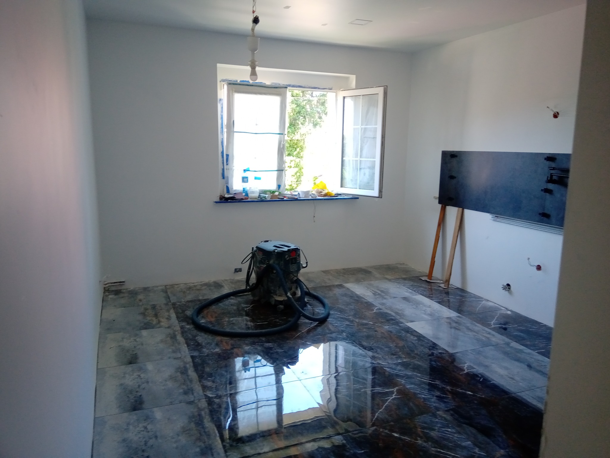 Wnętrze pokoju z nową, błyszczącą podłogą z ciemnych płytek imitujących marmur, na której stoi odkurzacz przemysłowy. Przy oknie folia ochronna. Ściany białe, przygotowane do malowania.