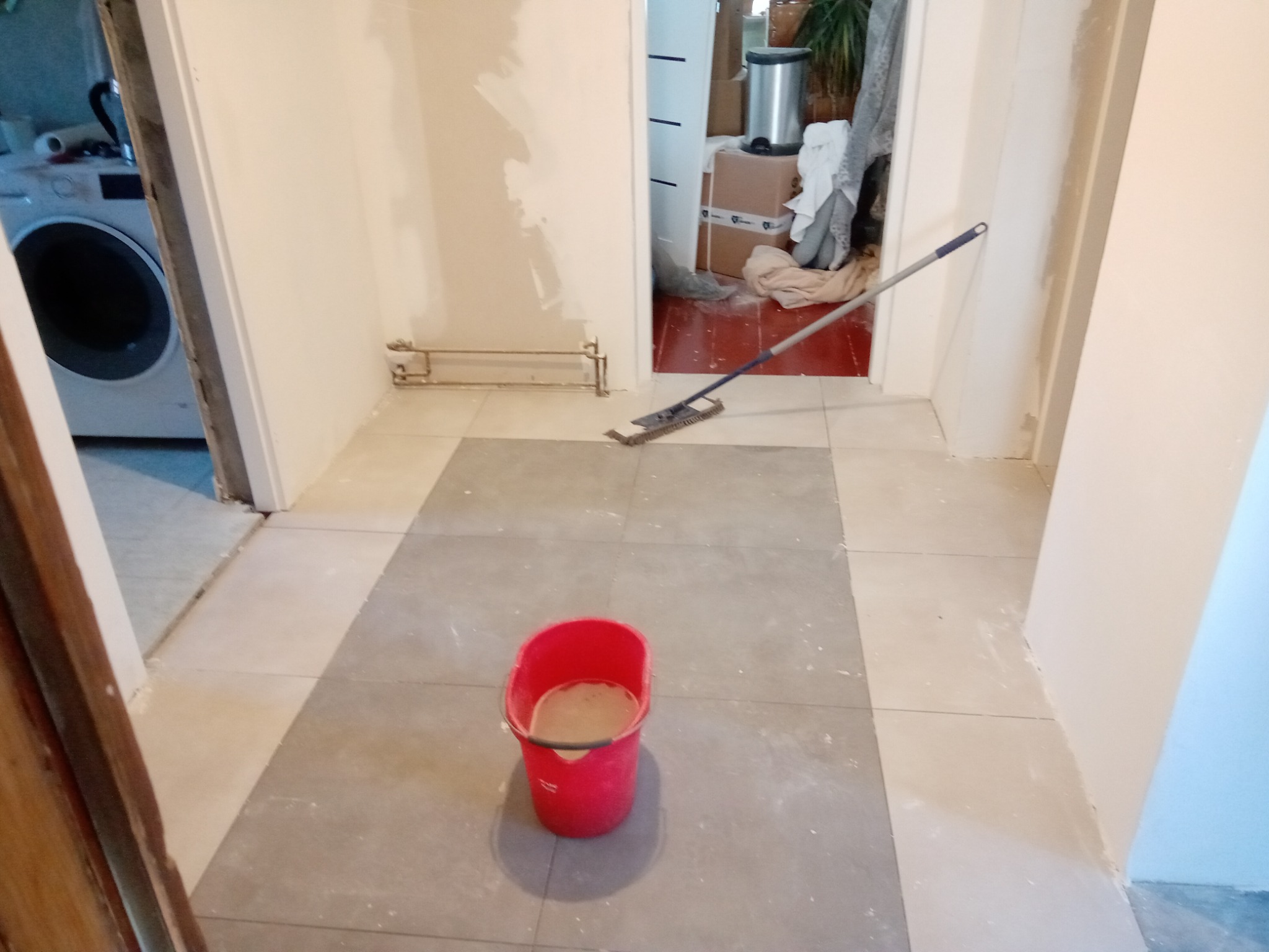 Wnętrze w trakcie prac wykończeniowych: świeżo położona podłoga z płytek w dwóch odcieniach szarości, czerwone wiadro z brudną wodą i mop leżący na podłodze, ściany po szpachlowaniu.
