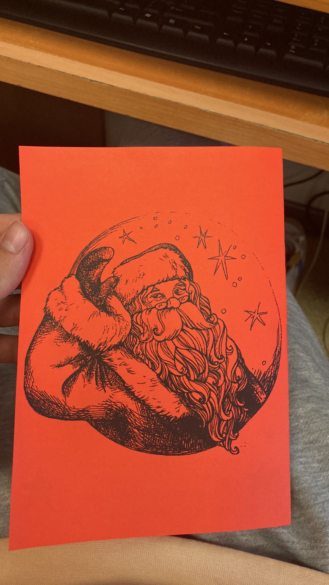 Czerwona kartka z ręcznie rysowanym portretem Świętego Mikołaja w czerni, otoczonego gwiazdami i kropkami, trzymana w dłoni na tle biurka z klawiaturą.