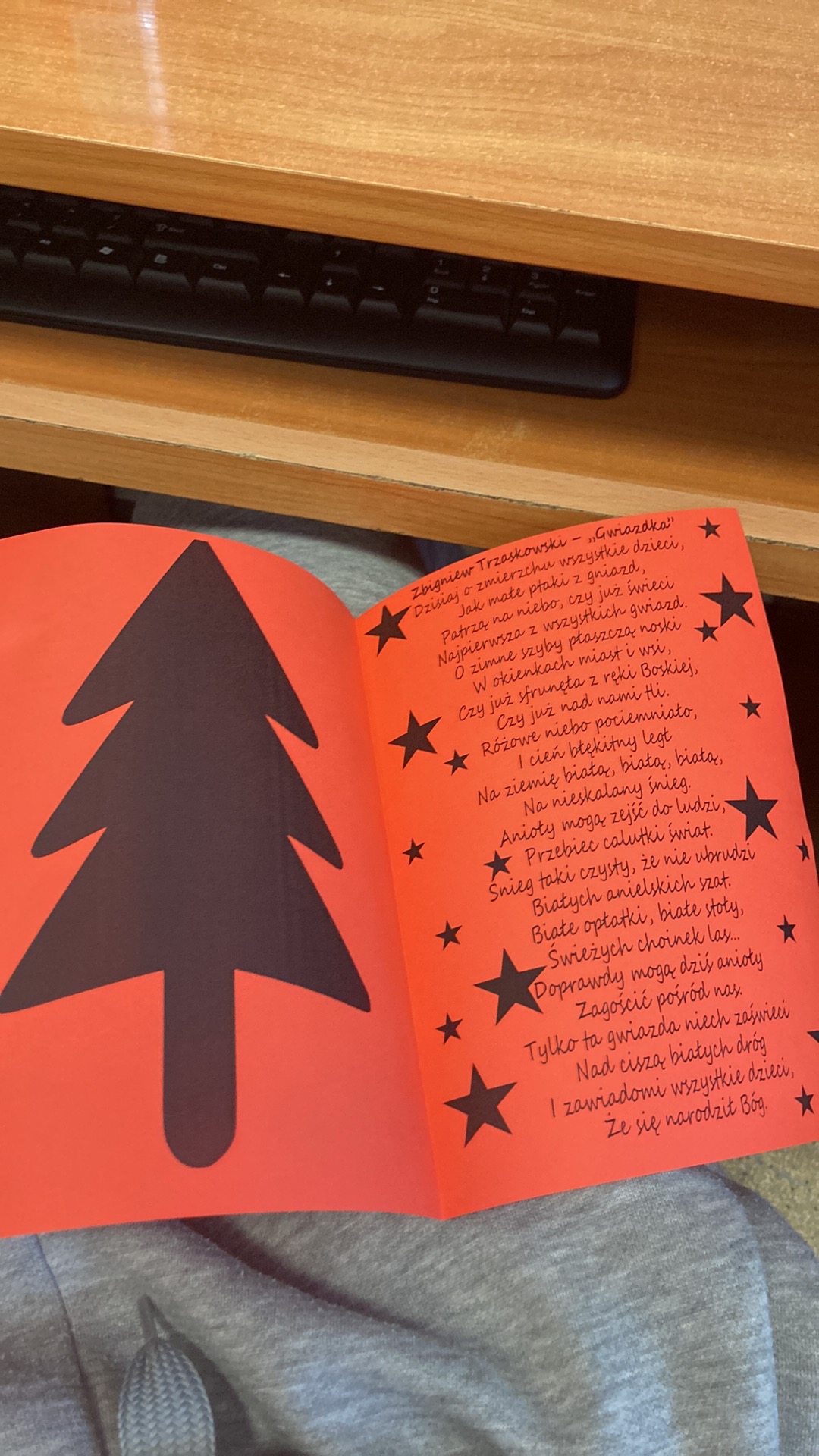 Otwarta, czerwona książka z czarnym rysunkiem choinki i gwiazd oraz wierszem Zbigniewa Trzaskowskiego 'Gwiazdka', leżąca na szarych spodniach, z widoczną klawiaturą w tle.