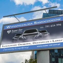Billboard reklamujący wypożyczalnię samochodów Super-Car z trzema samochodami na tle nieba i nowoczesnego budynku biurowego.