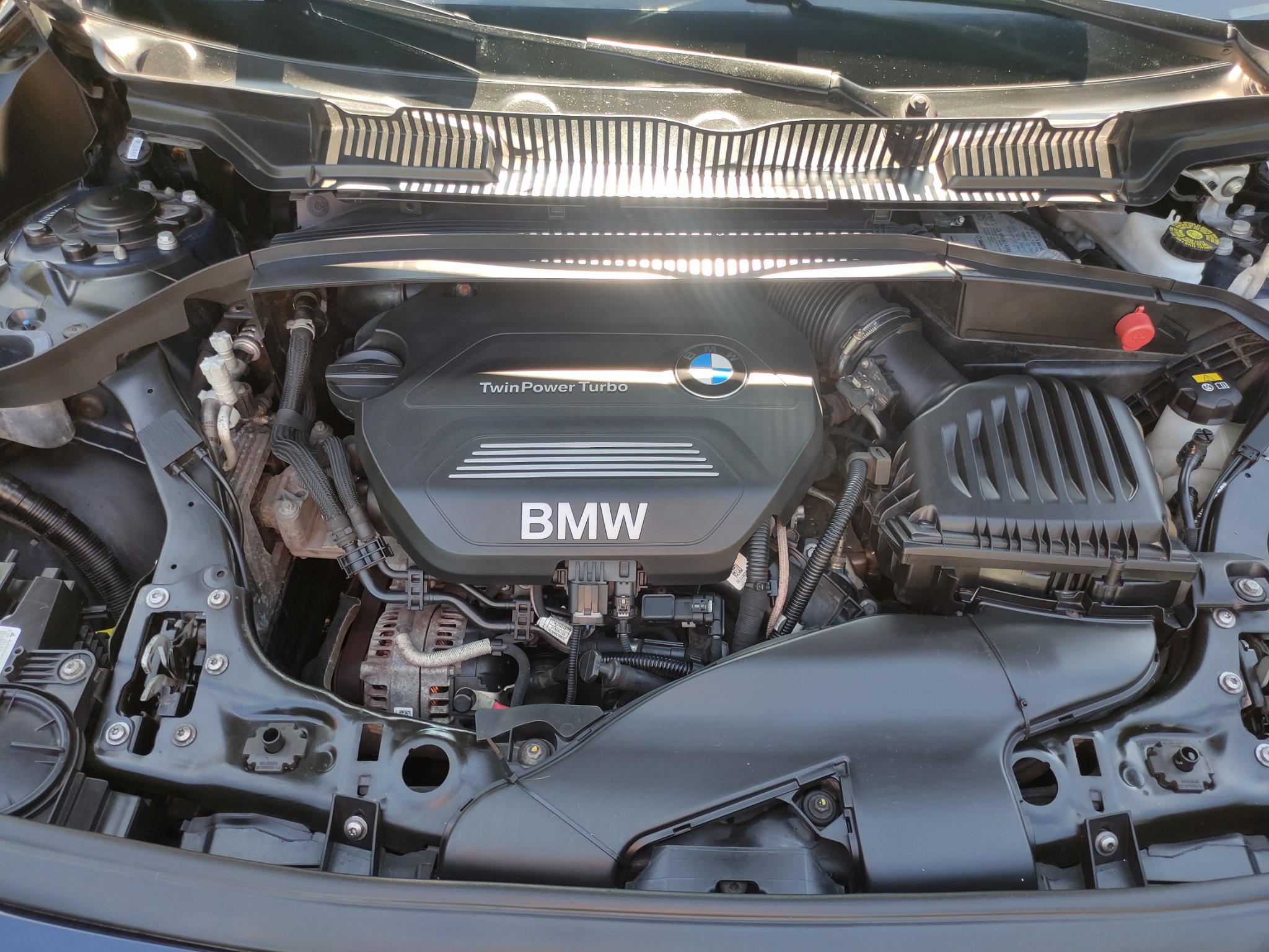 Otwarta maska samochodu marki BMW, widoczny silnik TwinPower Turbo z elementami osprzętu i przewodami, oświetlony promieniami słonecznymi.