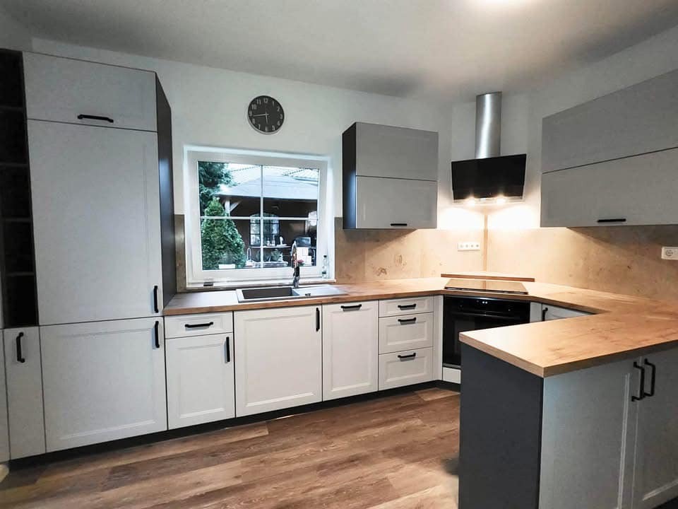 Nowoczesna kuchnia w odcieniach bieli i szarości z drewnianym blatem, czarnymi uchwytami i widokiem na ogród przez okno.