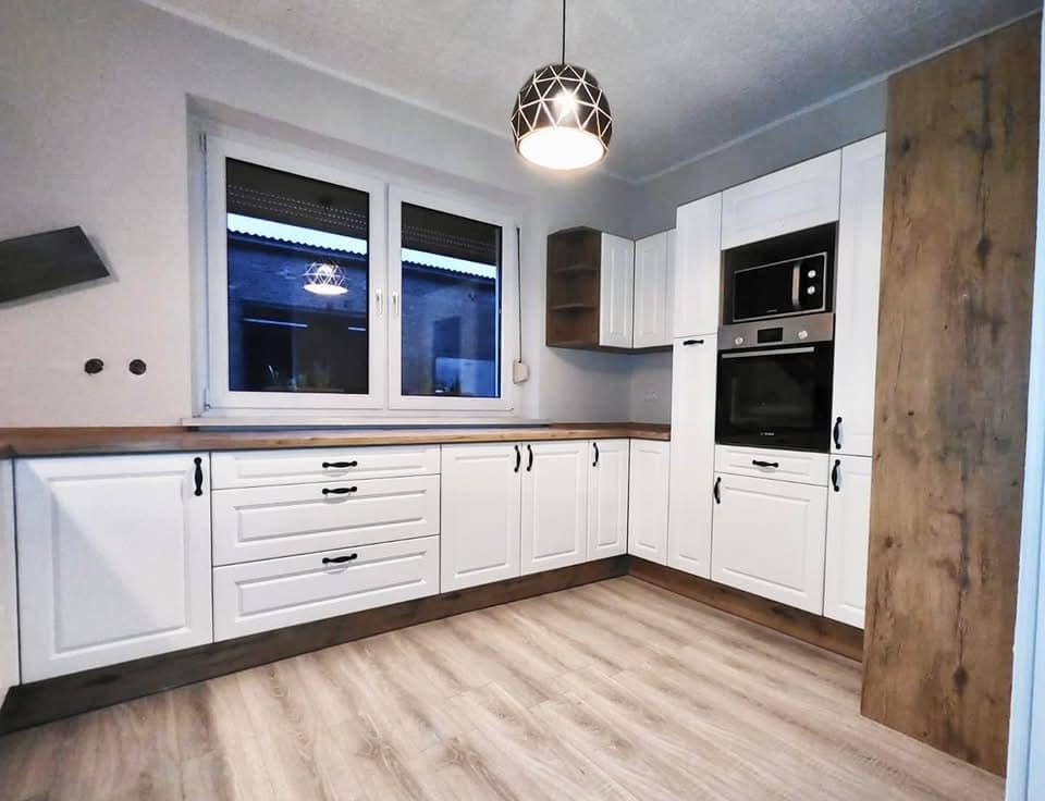 Jasna kuchnia w stylu rustykalnym z białymi szafkami, drewnianym blatem i podłogą, widok na okno z roletami i lampę wiszącą w geometrycznym kloszu.