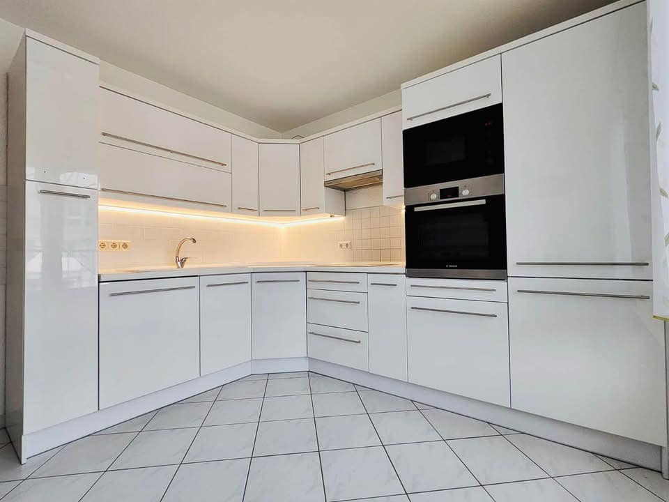 Jasna, nowoczesna kuchnia z białymi, lakierowanymi frontami, podświetleniem LED nad blatem i sprzętem AGD w zabudowie, widok na białą podłogę z płytek w szachownicę.