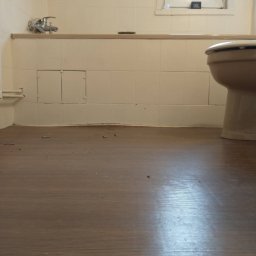 Kamilos Remontuj Z Pasją - Brązowa podłoga w łazience z widocznymi niedobitkami po remoncie, białe kafelki na ścianach, toaleta i okno z głośnikiem JBL na parapecie.