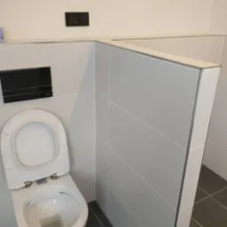 Nowoczesna toaleta wisząca z otwartą klapą, czarny panel spłuczki i szare płytki podłogowe, wykończona listwami aluminiowymi.