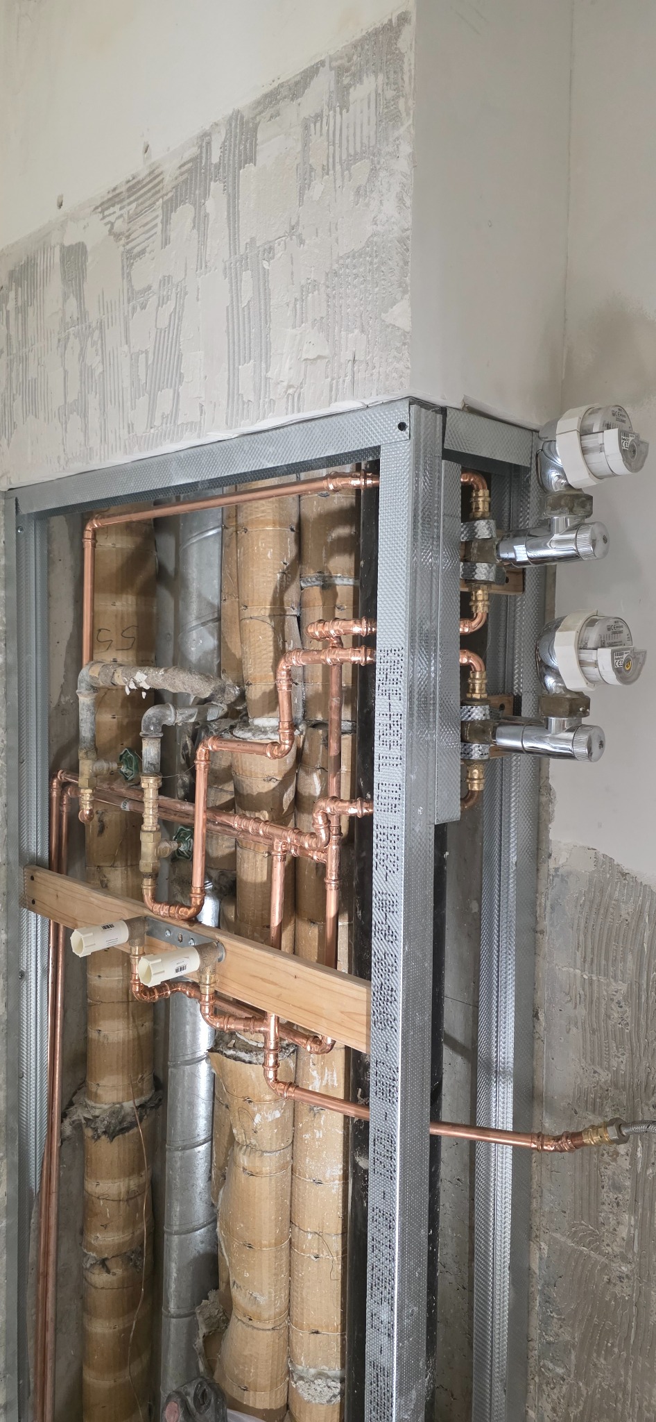 Miedziana instalacja wodna w trakcie montażu, widoczne rury, zawory i metalowa konstrukcja stelaża na tle surowej ściany w trakcie remontu.