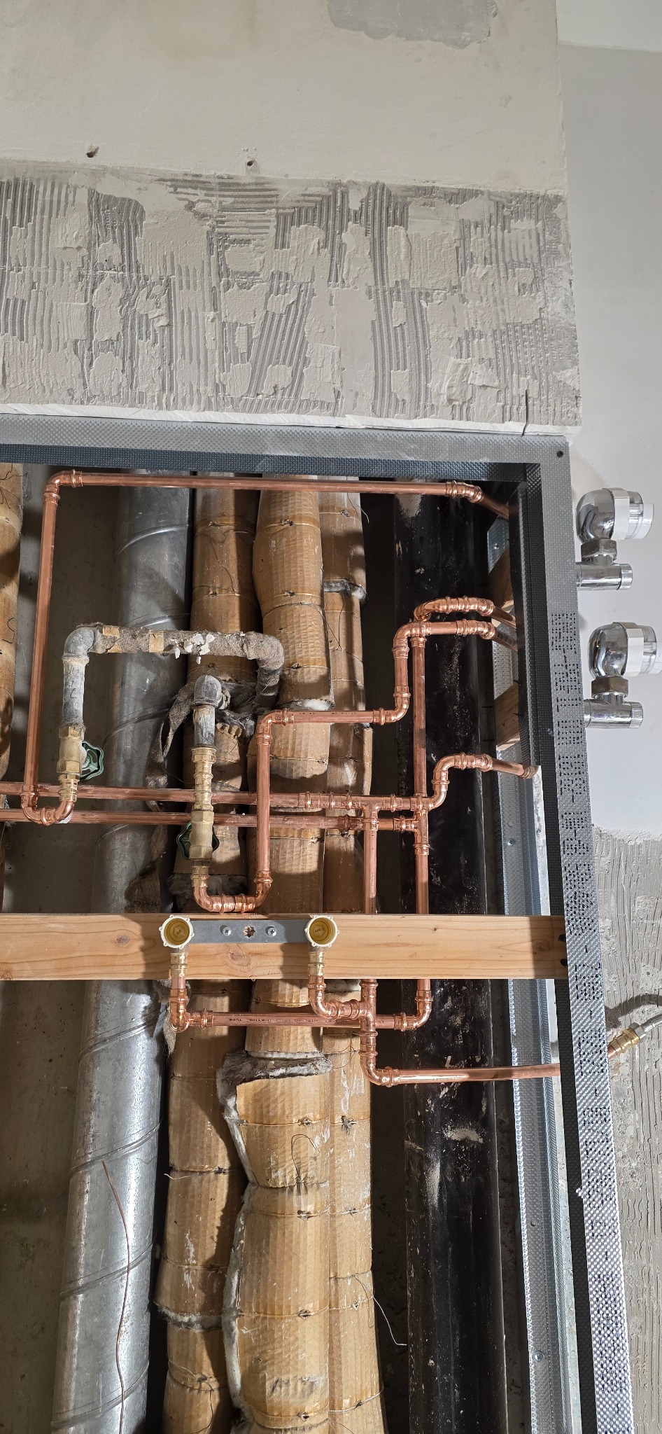 Miedziana instalacja wodna w trakcie montażu, widoczne rury, izolacja, stelaż i fragment ściany z resztkami tynku. Instalacja w ramie ze stelażu.