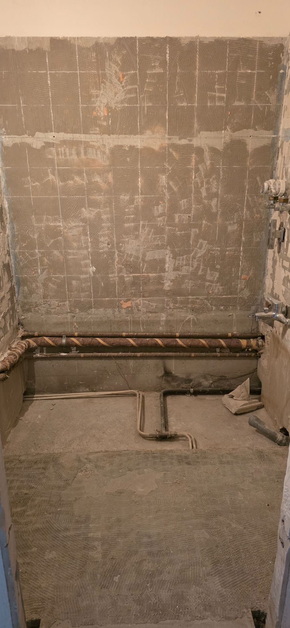 Przygotowana do remontu łazienka: ściany z resztkami kleju po płytkach, rury instalacyjne, surowa podłoga. Widok na wnętrze w trakcie prac remontowych.