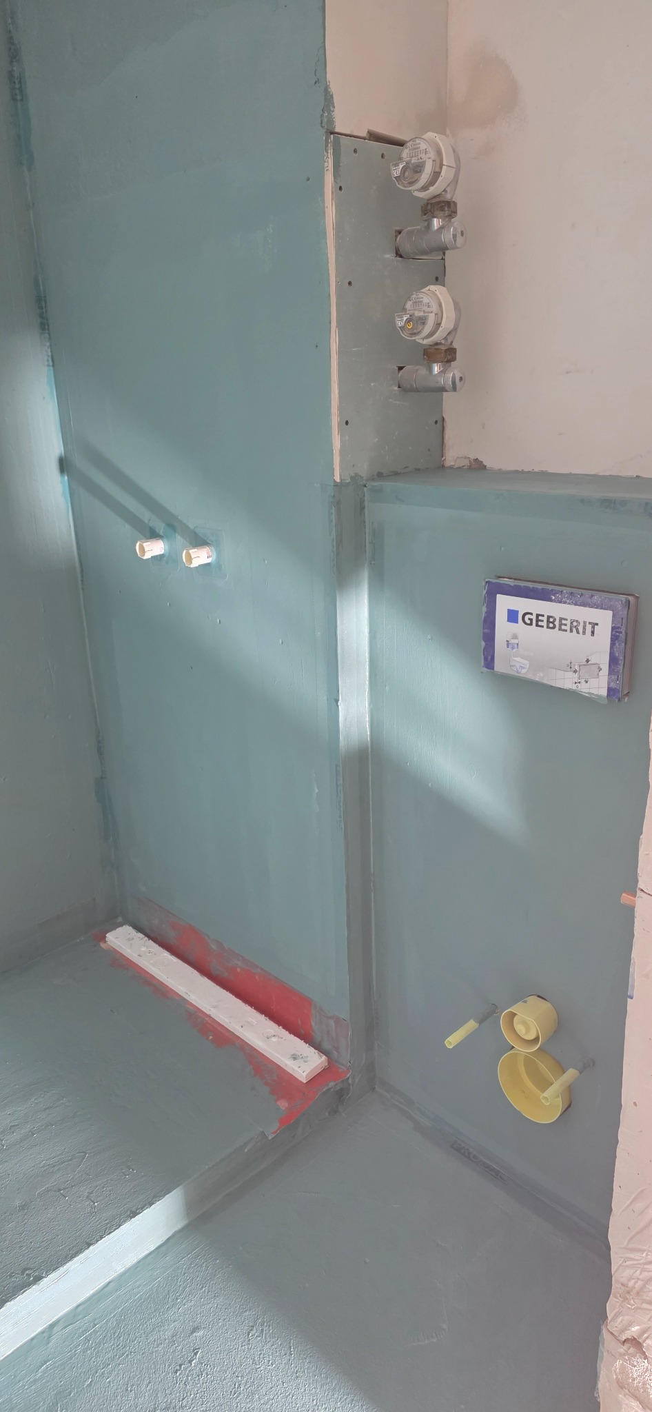 Hydroizolacja w rogu łazienki z zamontowanymi elementami instalacji wodnej i odpływem liniowym, przygotowana do dalszych prac wykończeniowych.