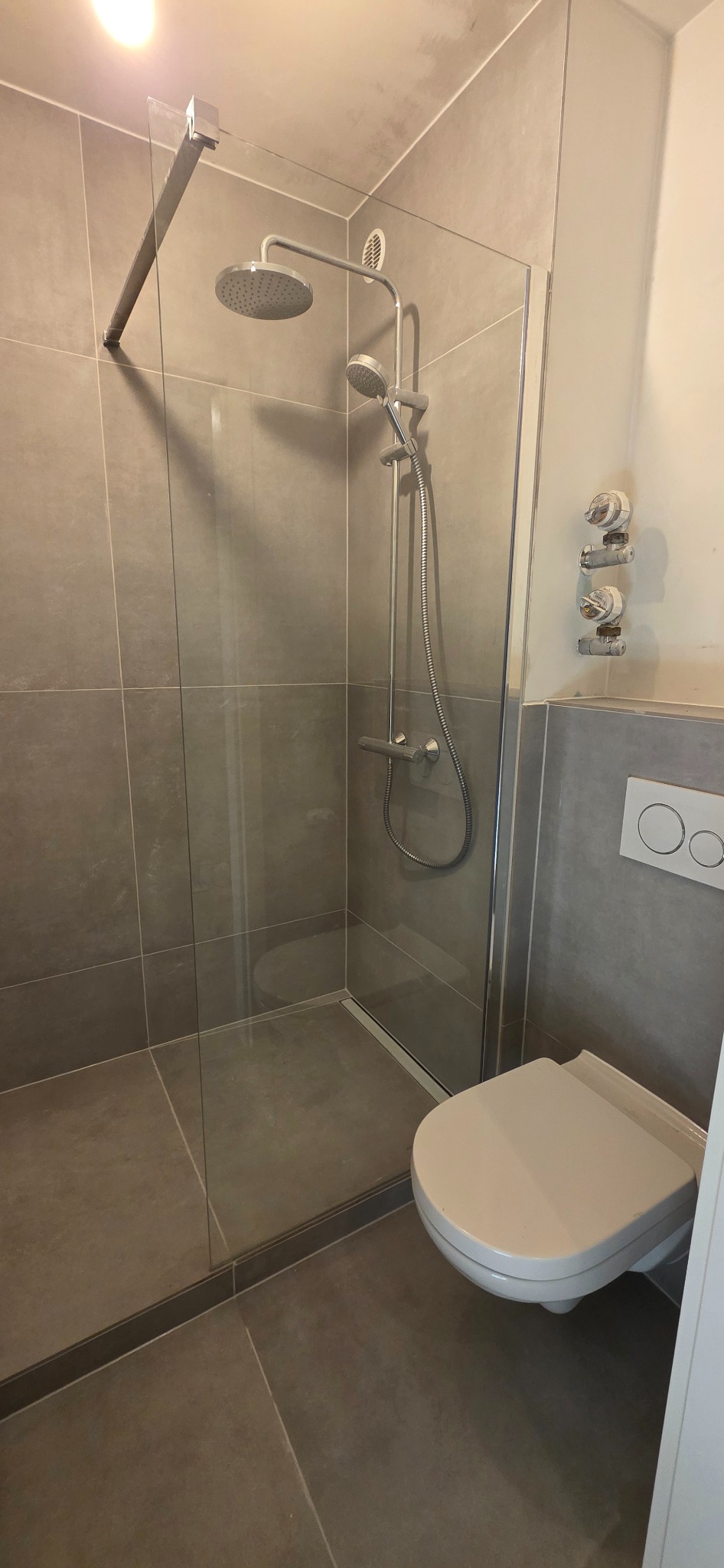 Nowoczesna kabina prysznicowa walk-in z deszczownicą i odpływem liniowym, wykończona szarą płytką wielkoformatową. Widoczna toaleta podwieszana.