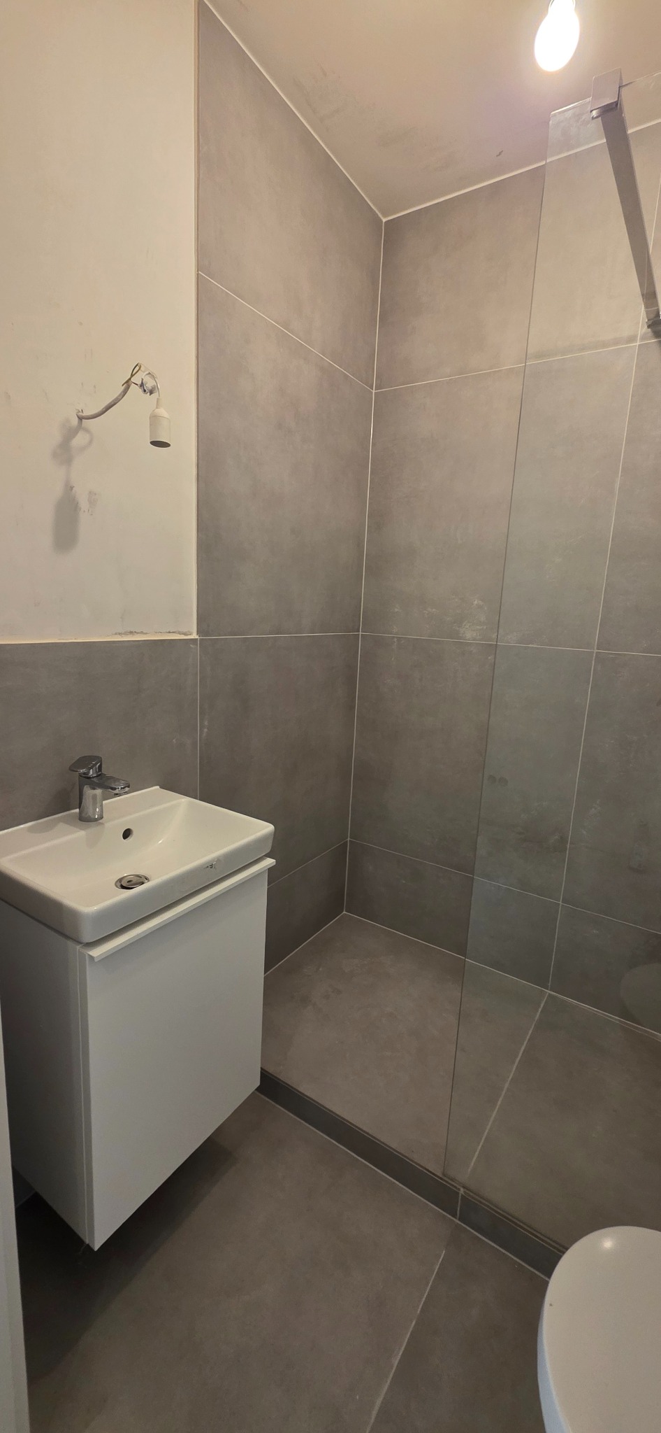 Nowoczesna łazienka z szarymi płytkami, białą szafką pod umywalką i szklaną kabiną prysznicową. Minimalistyczny design, surowy wystrój, widoczny fragment toalety.