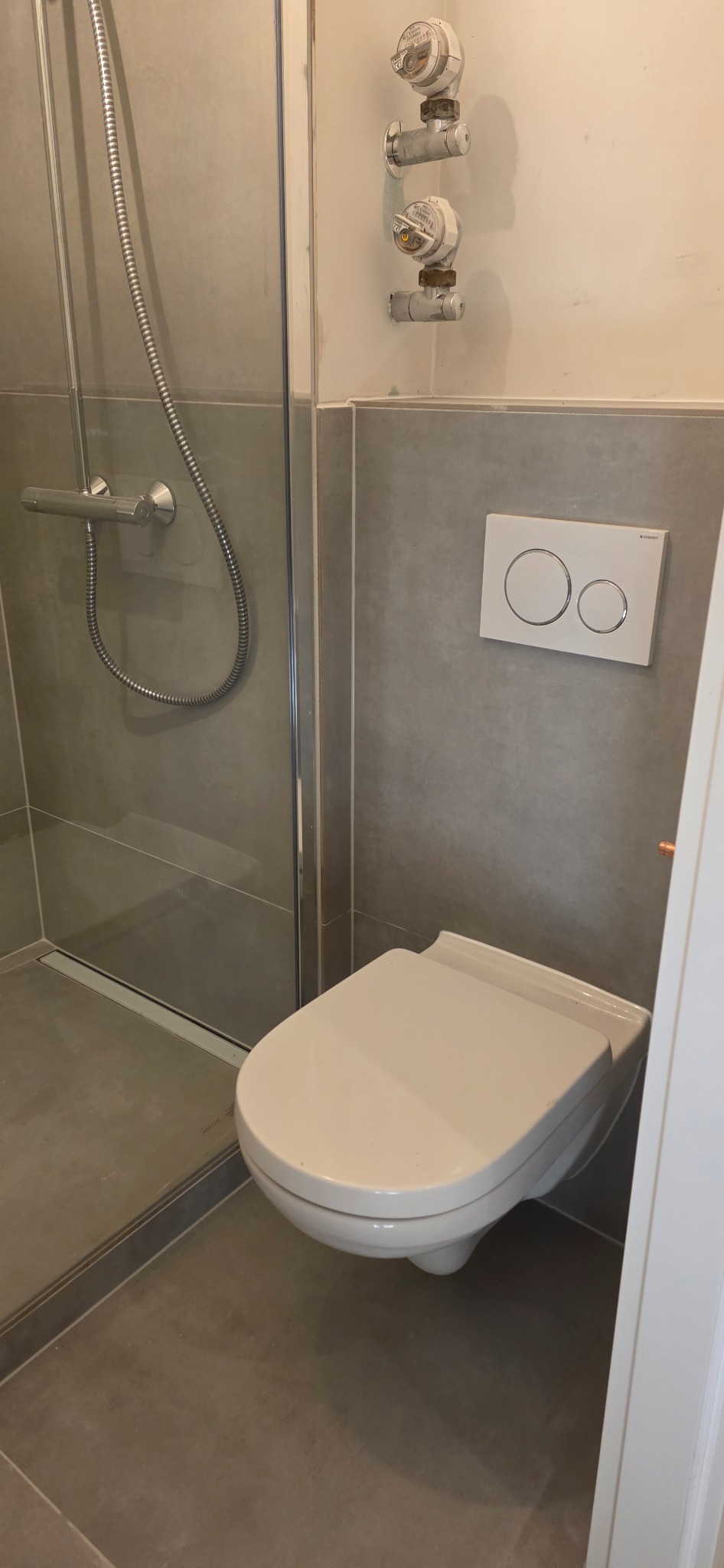 Nowoczesna łazienka po remoncie: szara glazura, podwieszana toaleta z przyciskiem Geberit, kabina prysznicowa ze srebrnym panelem i odpływem liniowym.