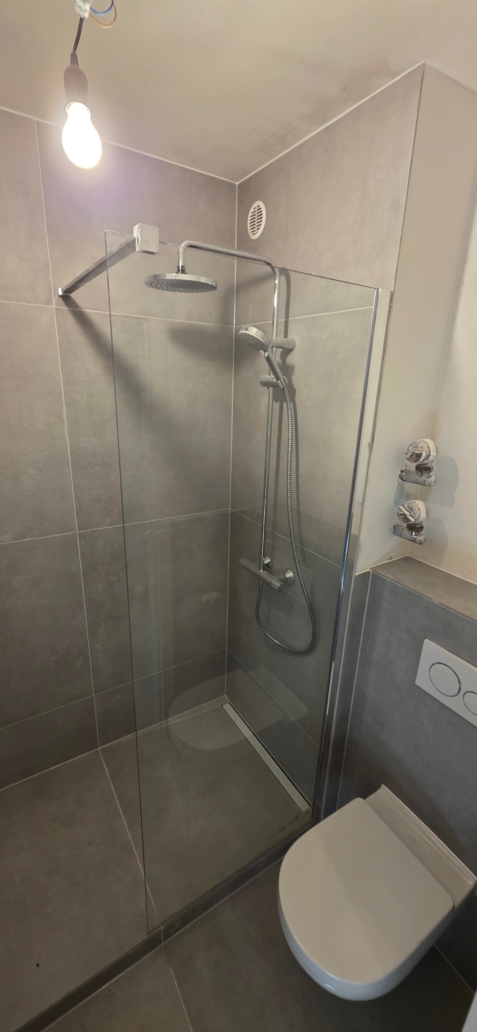 Szara łazienka z kabiną prysznicową typu walk-in, widoczny fragment toalety. Minimalistyczny design, płytki imitujące beton. Żarówka na kablu zamiast oświetlenia.