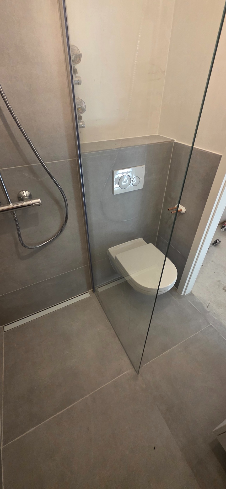 Nowoczesna łazienka z kabiną prysznicową typu walk-in i podwieszaną toaletą. Szare płytki, minimalistyczny design, widoczny odpływ liniowy i bateria prysznicowa.