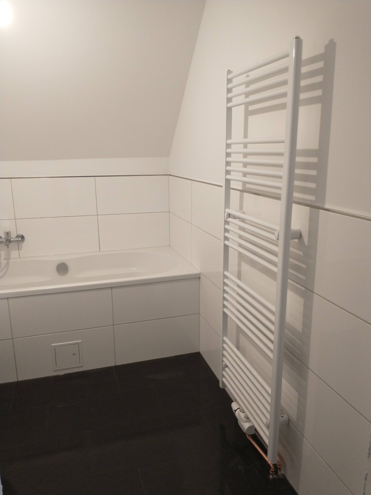 Biała łazienka z wanną obłożoną białymi płytkami, ciemną podłogą i grzejnikiem drabinkowym na ścianie.