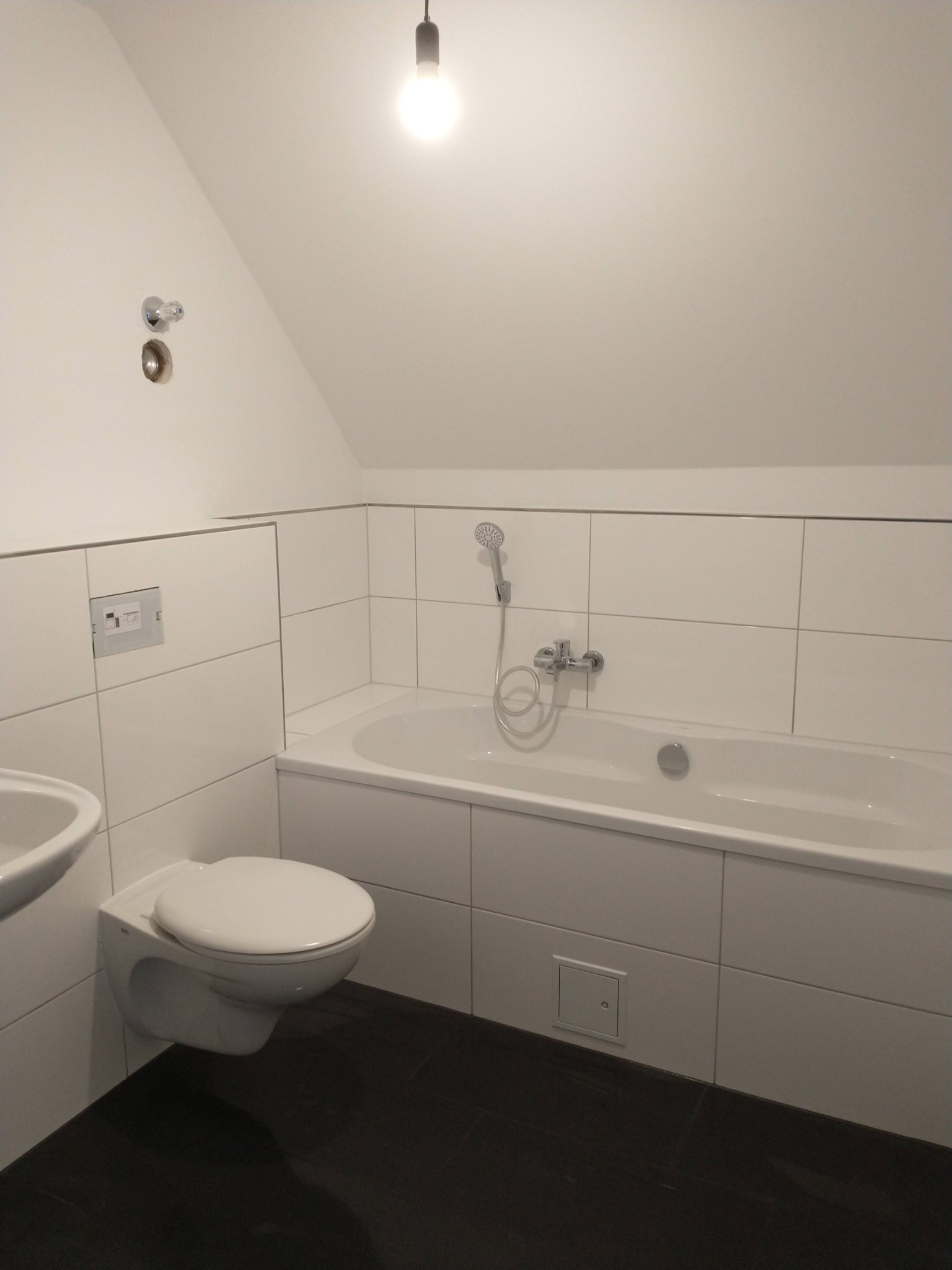 Nowoczesna łazienka na poddaszu z białą armaturą, wanną obudowaną płytkami i ciemną podłogą, oświetlona pojedynczą żarówką.