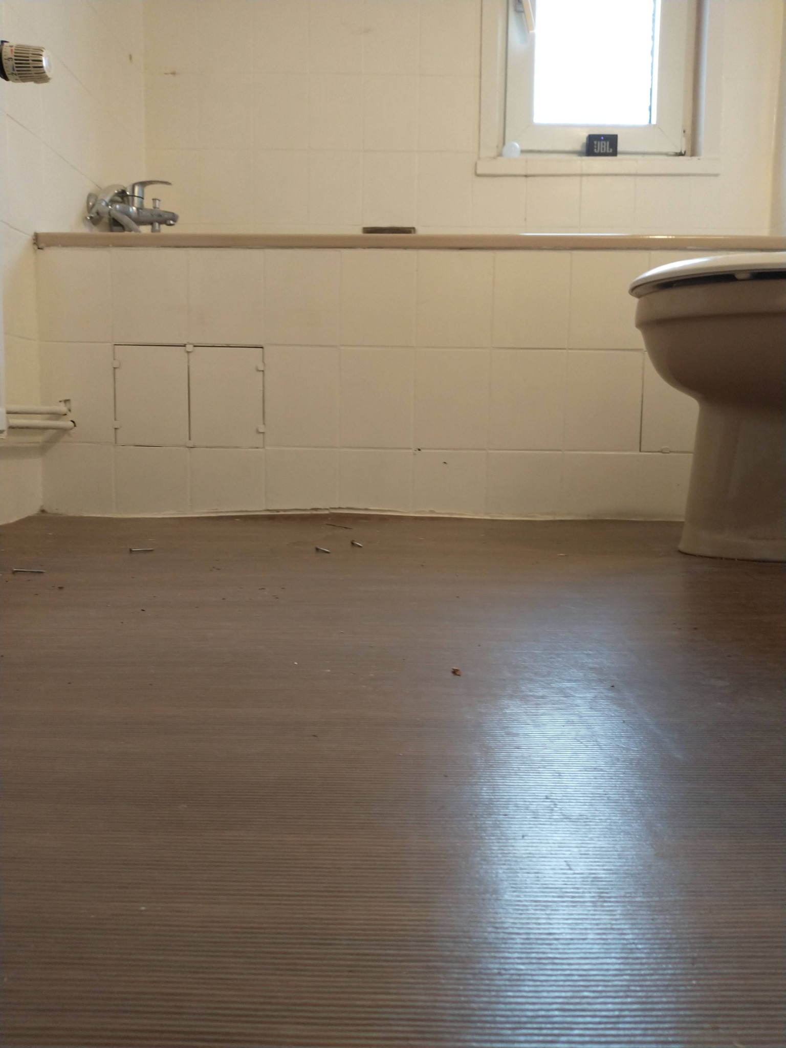 Brązowa podłoga w łazience z widocznymi niedobitkami po remoncie, białe kafelki na ścianach, toaleta i okno z głośnikiem JBL na parapecie.