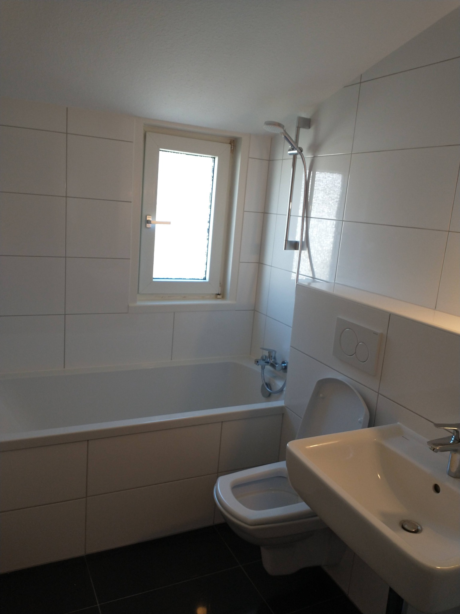 Biała łazienka z wanną, toaletą i umywalką, wykończona białymi płytkami i czarną podłogą, z oknem w białej ramie.