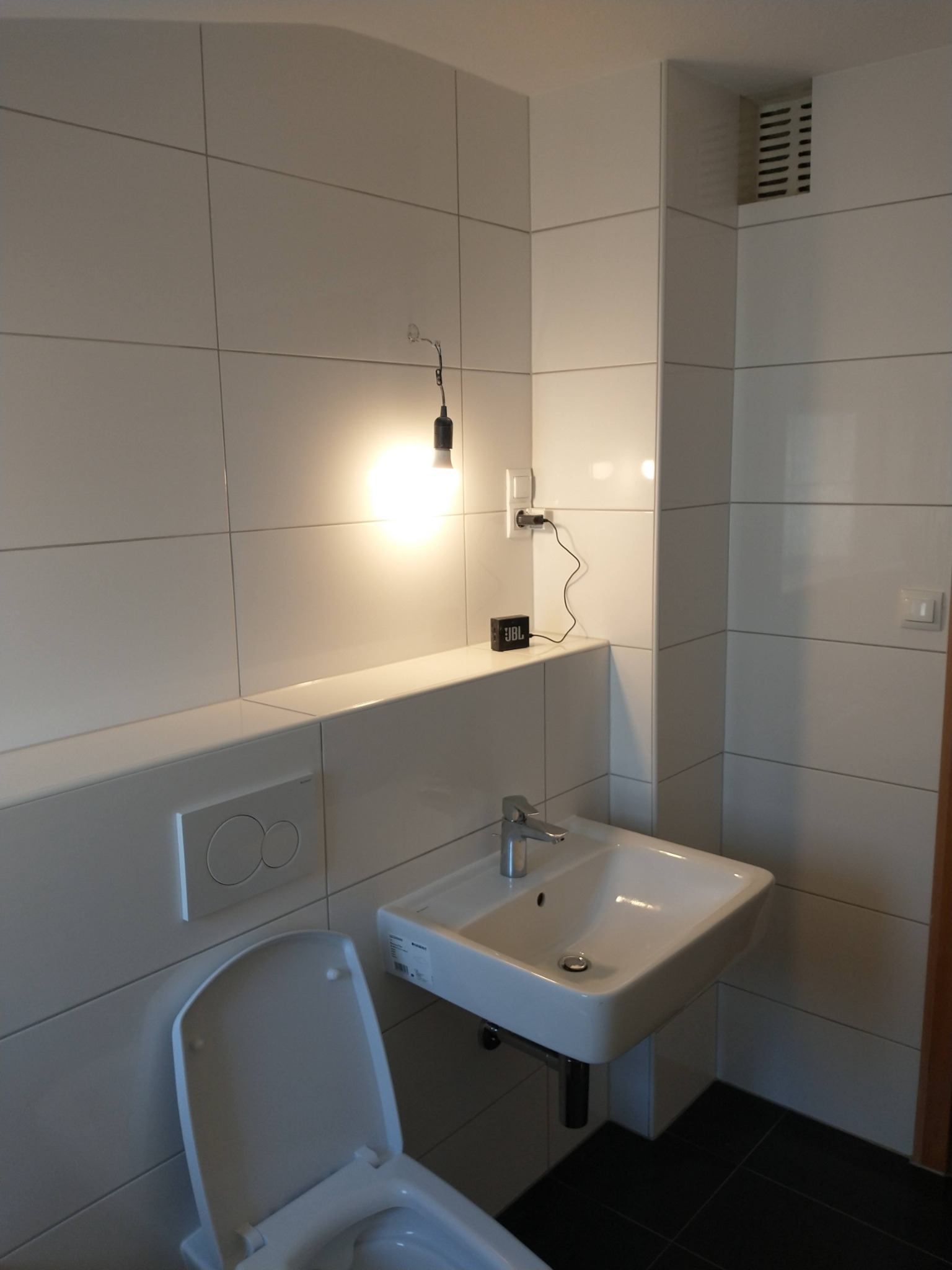 Nowoczesna łazienka z białymi płytkami, wiszącą umywalką, toaletą i minimalistycznym oświetleniem z żarówką na kablu oraz głośnikiem JBL na półce.