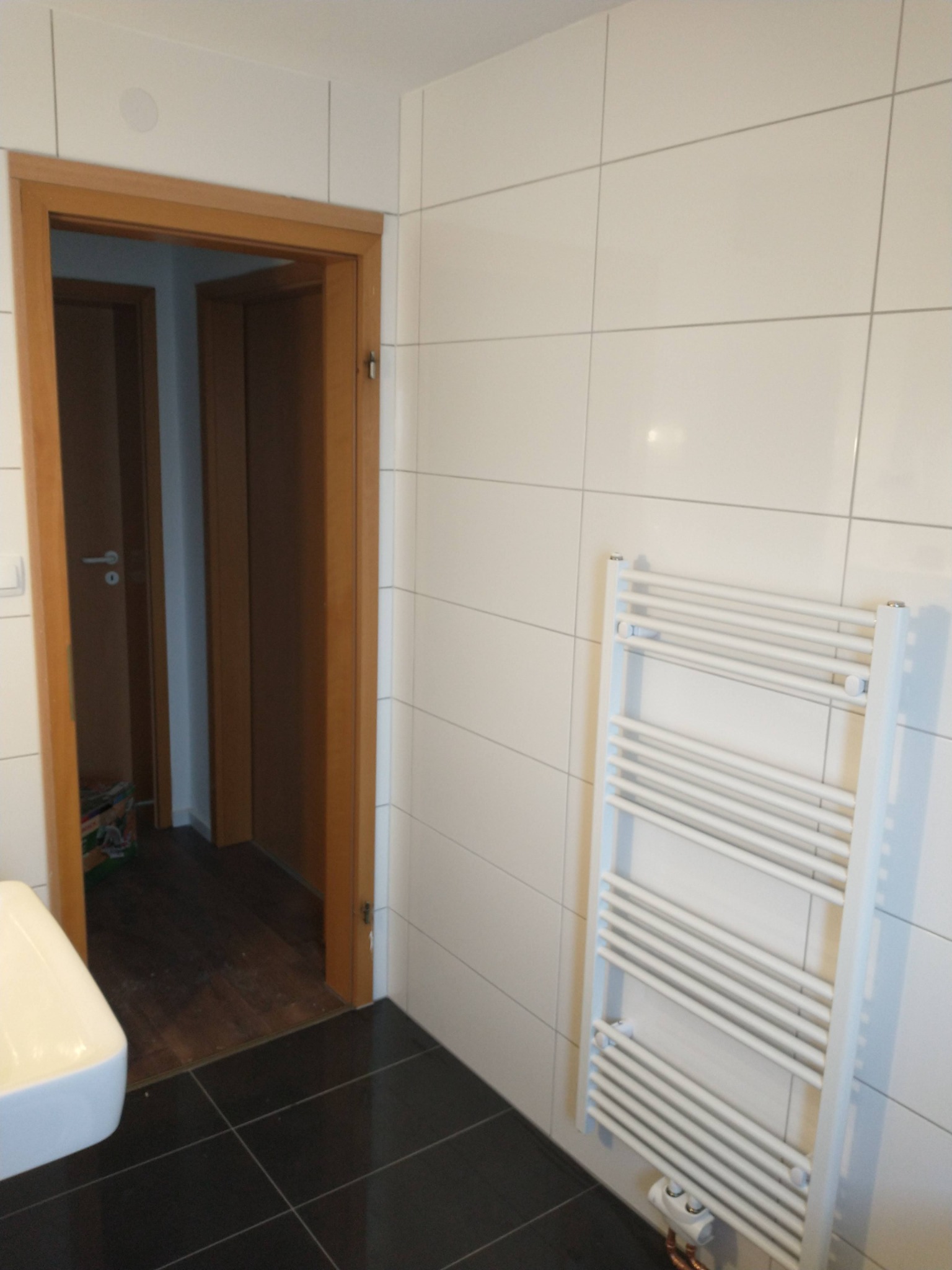 Biała łazienka z czarną podłogą, białe płytki na ścianach, grzejnik drabinkowy i drewniane obramowanie drzwi do innego pomieszczenia z drewnianą podłogą.