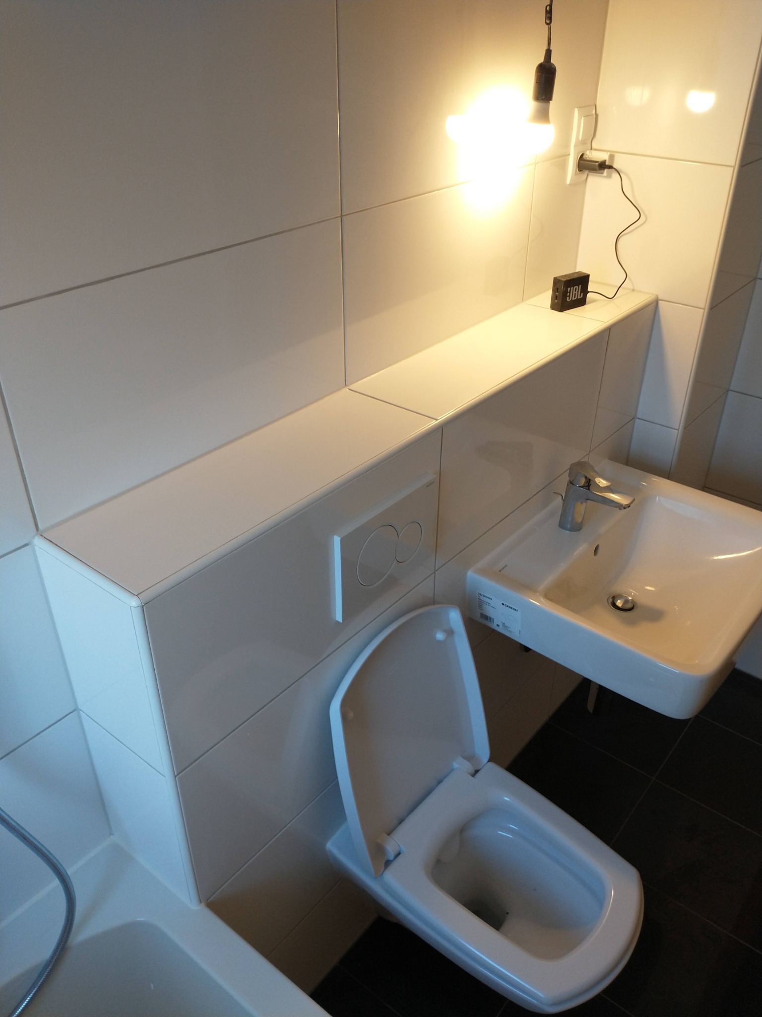 Nowoczesna łazienka z białymi płytkami, podwieszaną toaletą z otwartą deską, umywalką nablatową, czarną podłogą i wiszącą lampą z żarówką Edison oraz głośnikiem JBL na półce.