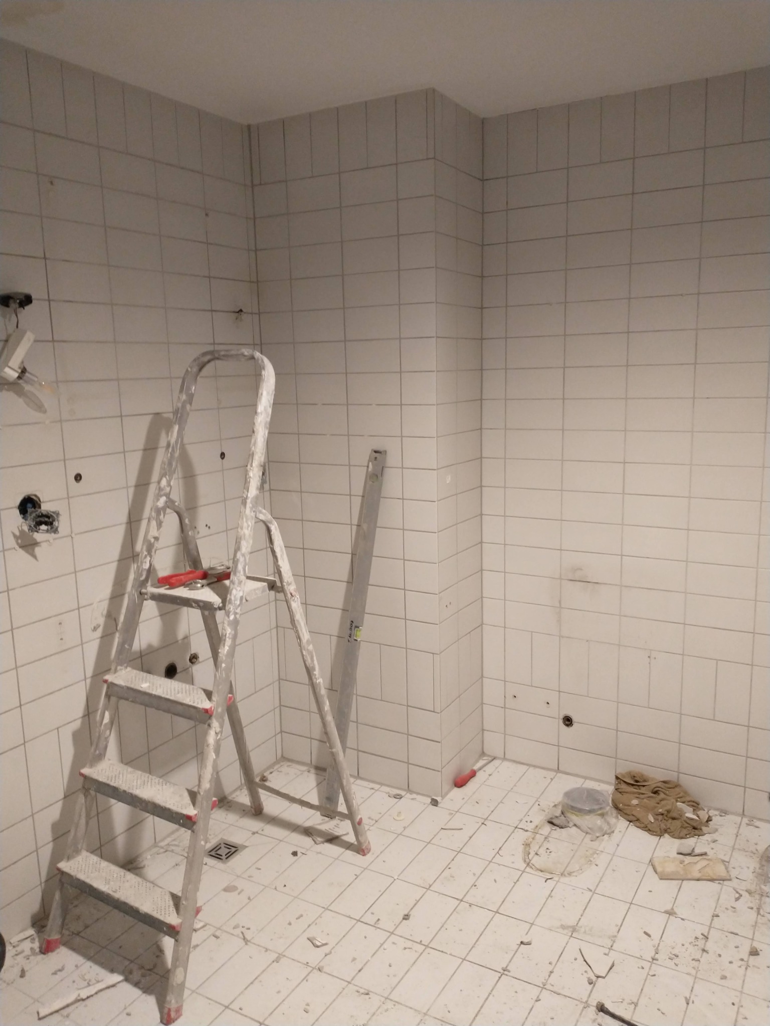 Remont łazienki w trakcie: białe płytki ścienne i podłogowe, aluminiowa drabina z narzędziami, poziomica oparta o ścianę, widoczne ślady po wierceniu w ścianach, gruz na podłodze.