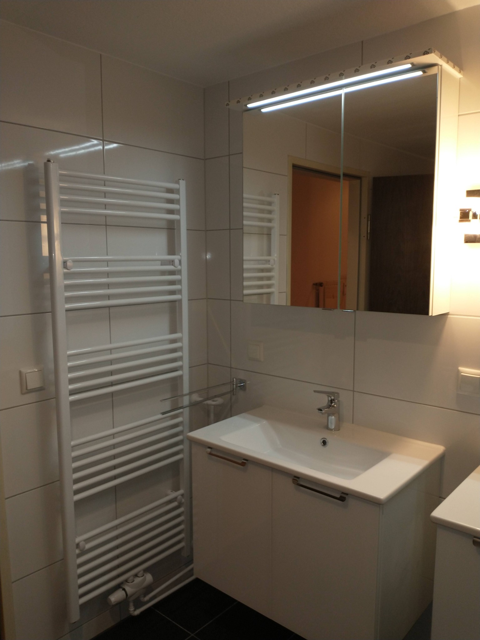 Biała łazienka w Dąbrowie Górniczej z grzejnikiem drabinkowym, umywalką z szafką i lustrem z oświetleniem LED.