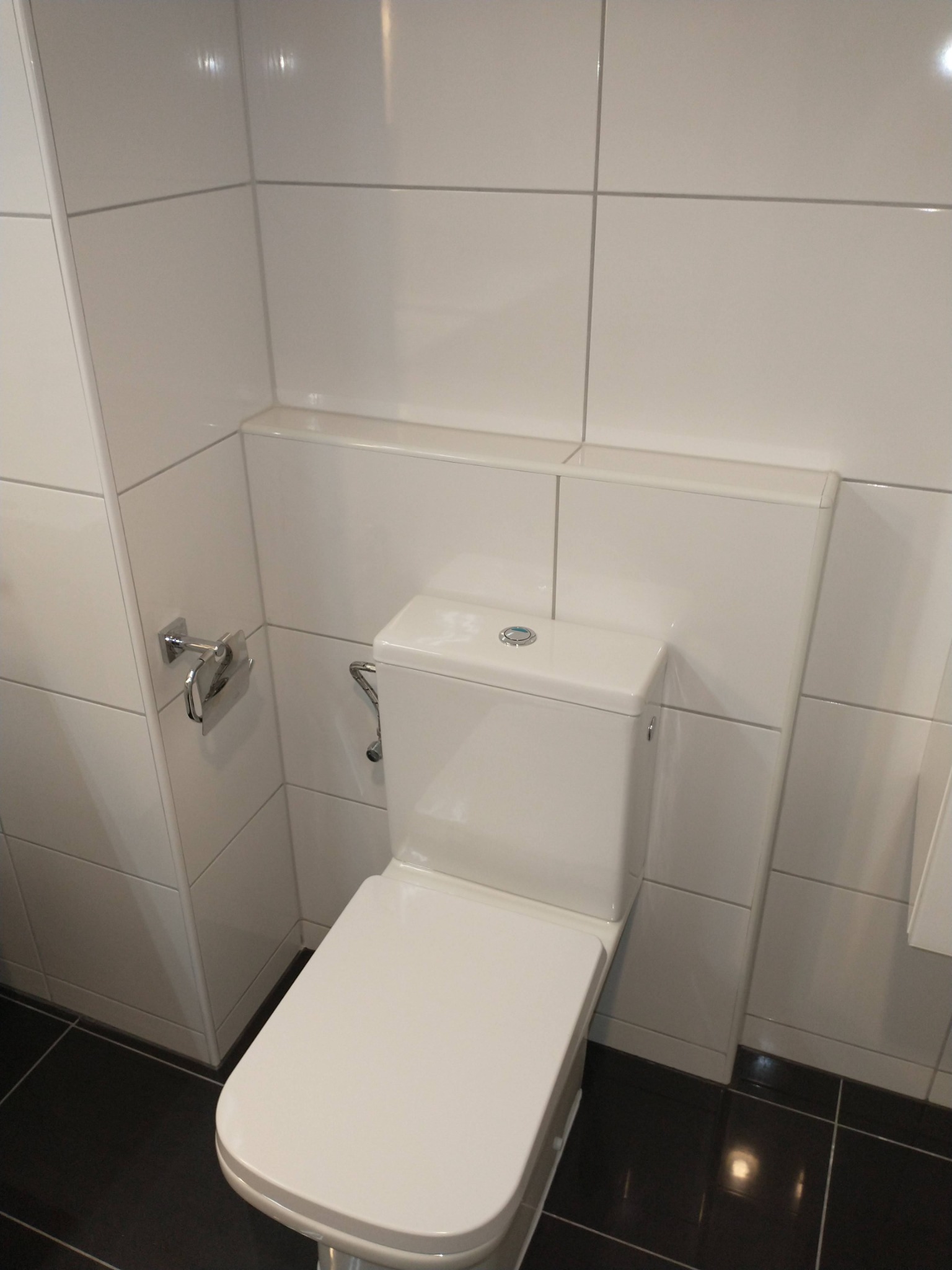 Nowoczesna toaleta z białą ceramiką, obudowana białymi płytkami, na podłodze ciemne płytki, widoczny uchwyt na papier toaletowy.