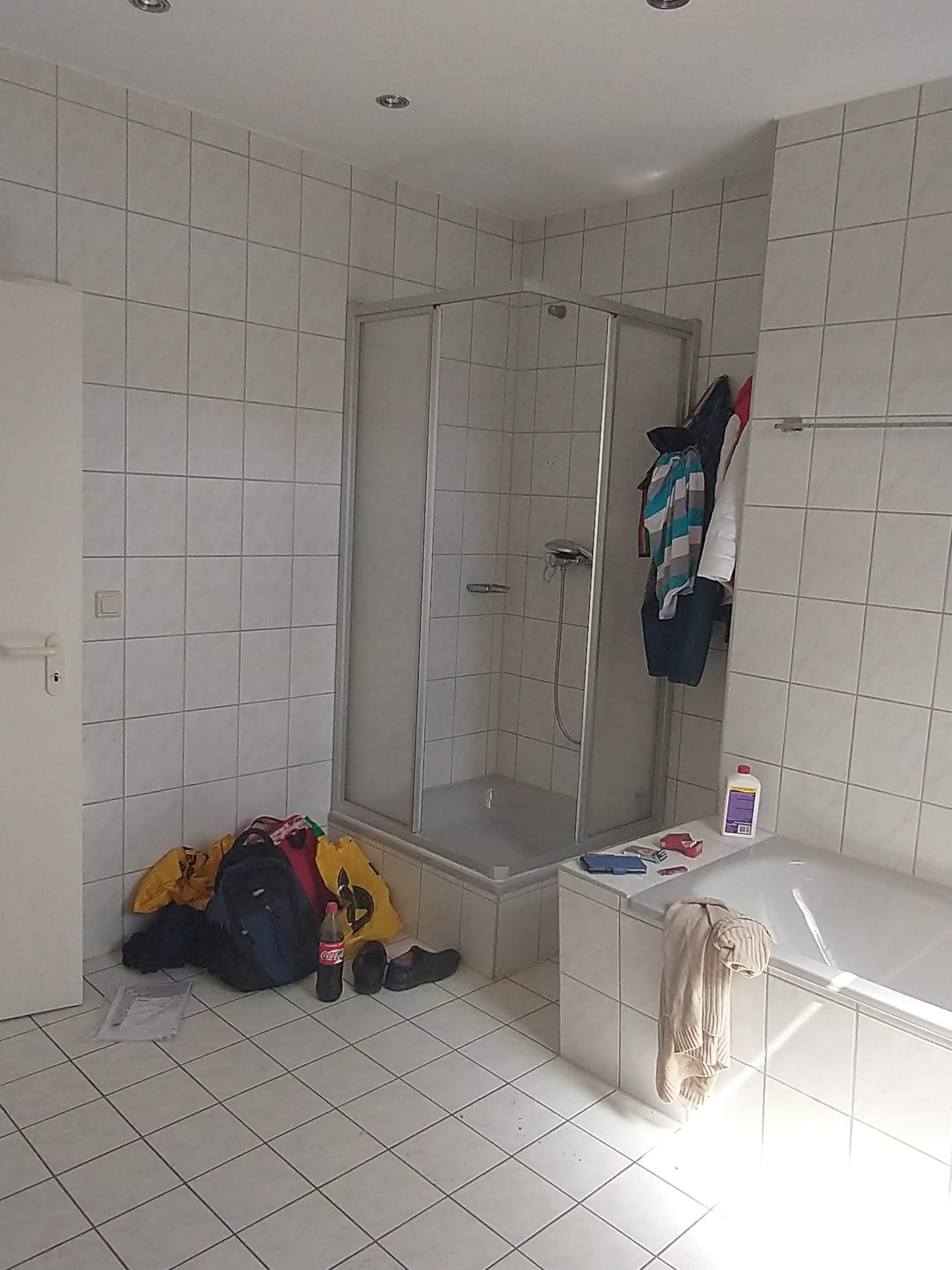 Łazienka z białymi płytkami, kabiną prysznicową i wanną; na podłodze i wannie porozrzucane torby, ubrania i kosmetyki, sugerujące remont lub wykończenie.