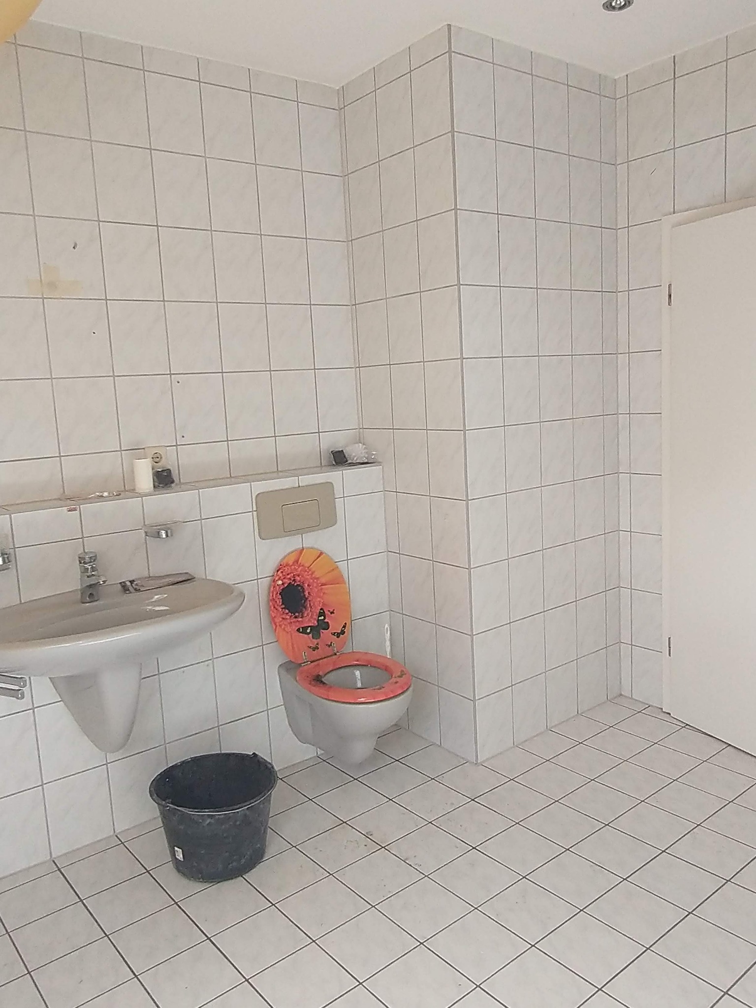 Łazienka przed remontem: białe płytki na ścianach i podłodze, umywalka z baterią, toaleta z pomarańczową deską w kwiaty, czarne wiadro na podłodze.