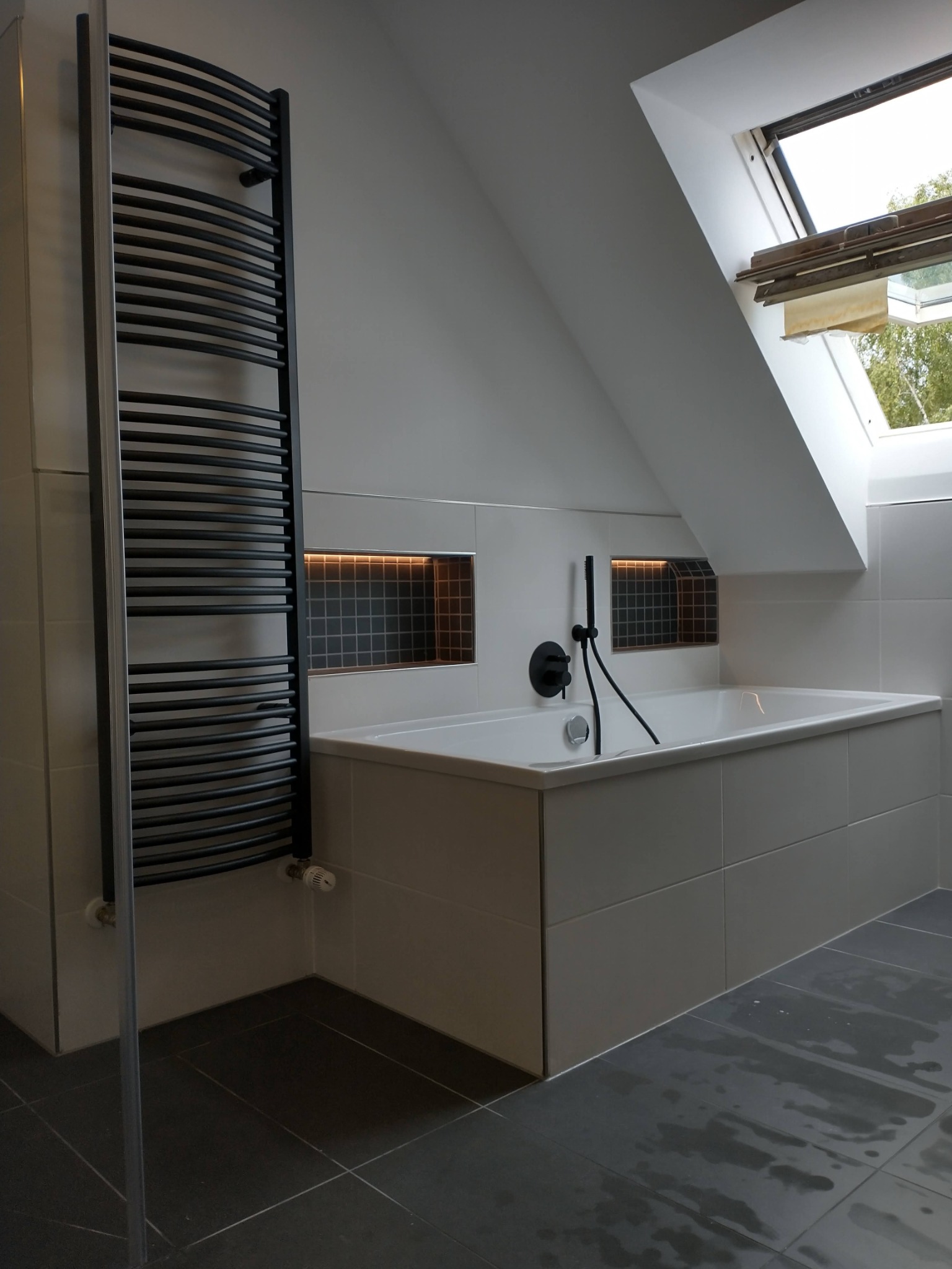 Nowoczesna łazienka na poddaszu z białą obudowaną wanną, czarną armaturą, grzejnikiem drabinkowym i oknem dachowym, wykończona szarą płytką podłogową i białymi płytkami ściennymi z wnękami z mozaiką.